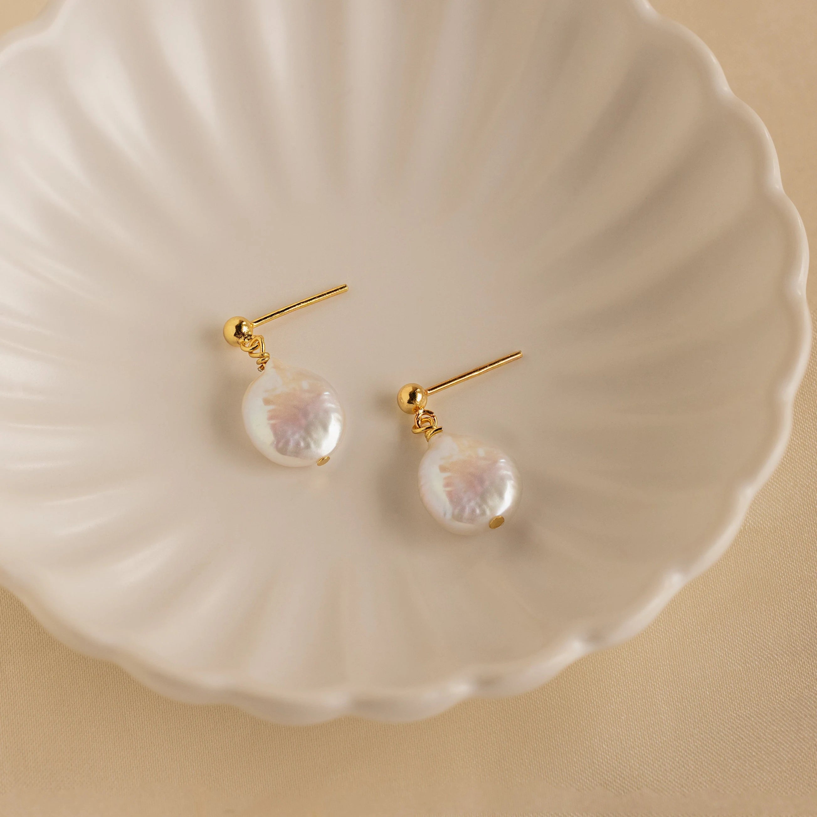 Round Pearl Drop Earrings featuring gold studs and iridescent round baroque pearls displayed on a white scalloped dish—an elegant accessory that brings timeless charm to any look.