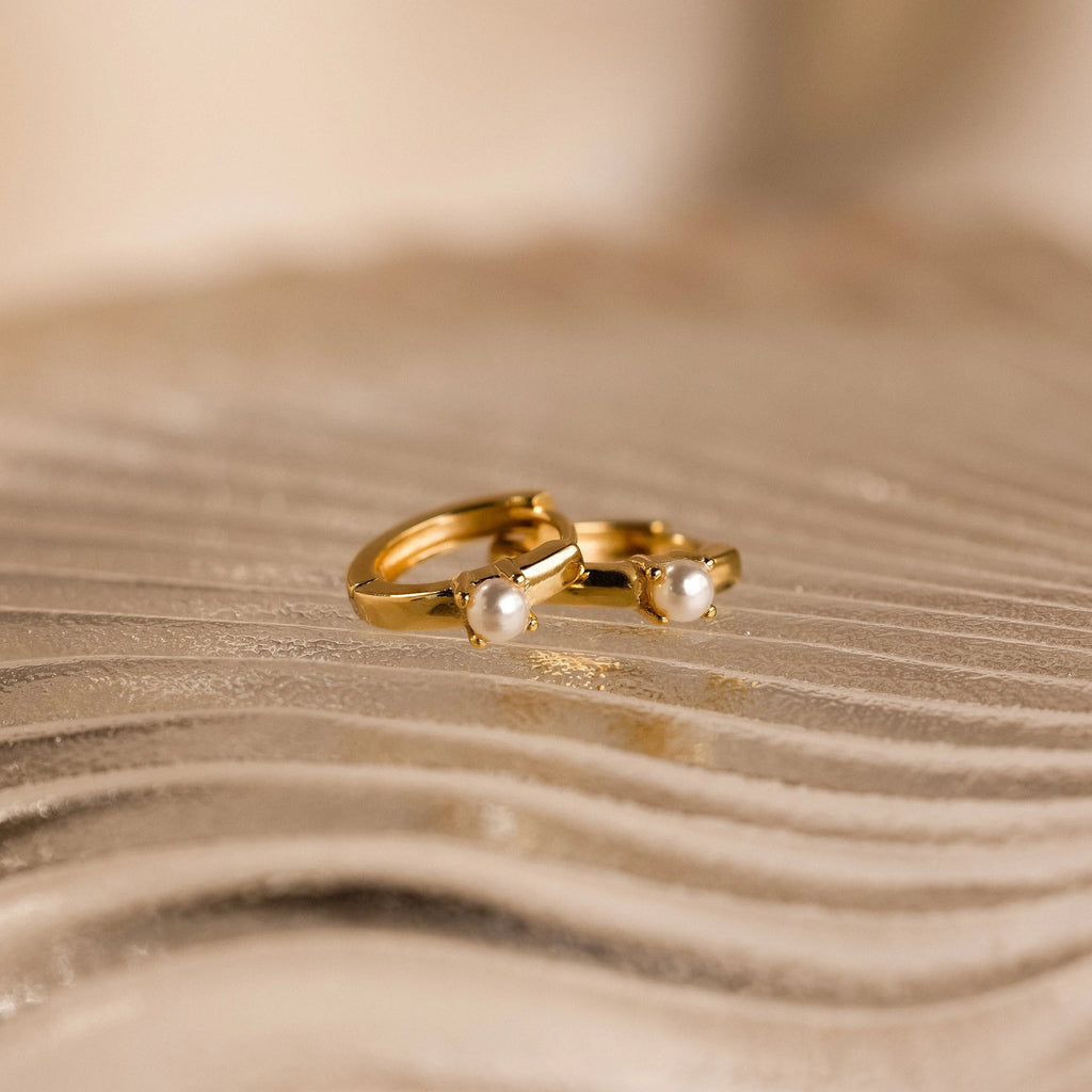 The Tiny Pearl Huggies, featuring two delicate gold hoops adorned with small pearls, rest elegantly on a textured, wavy beige surface.