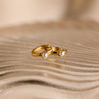 The Tiny Pearl Huggies, featuring two delicate gold hoops adorned with small pearls, rest elegantly on a textured, wavy beige surface.