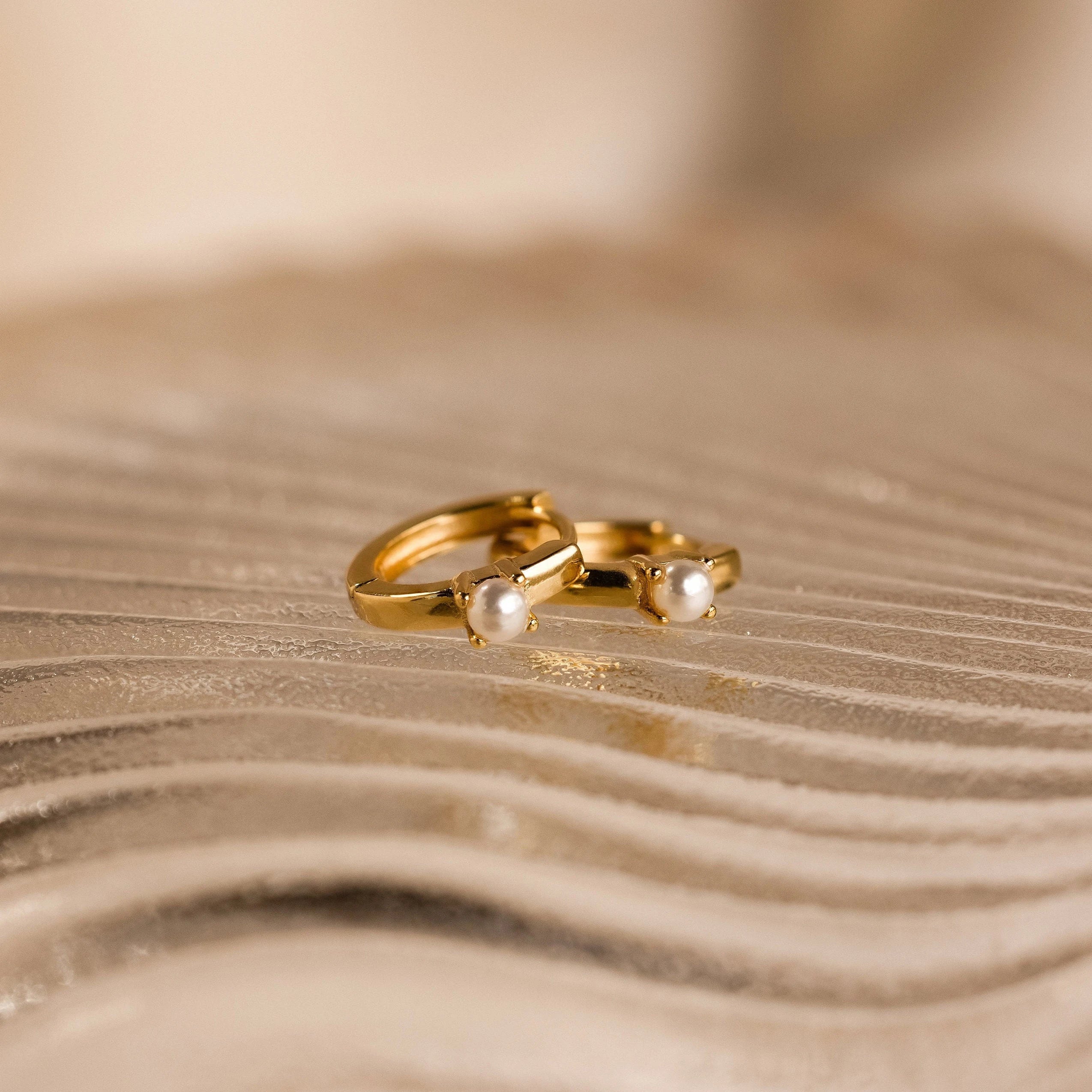 The Tiny Pearl Huggies, featuring two delicate gold hoops adorned with small pearls, rest elegantly on a textured, wavy beige surface.