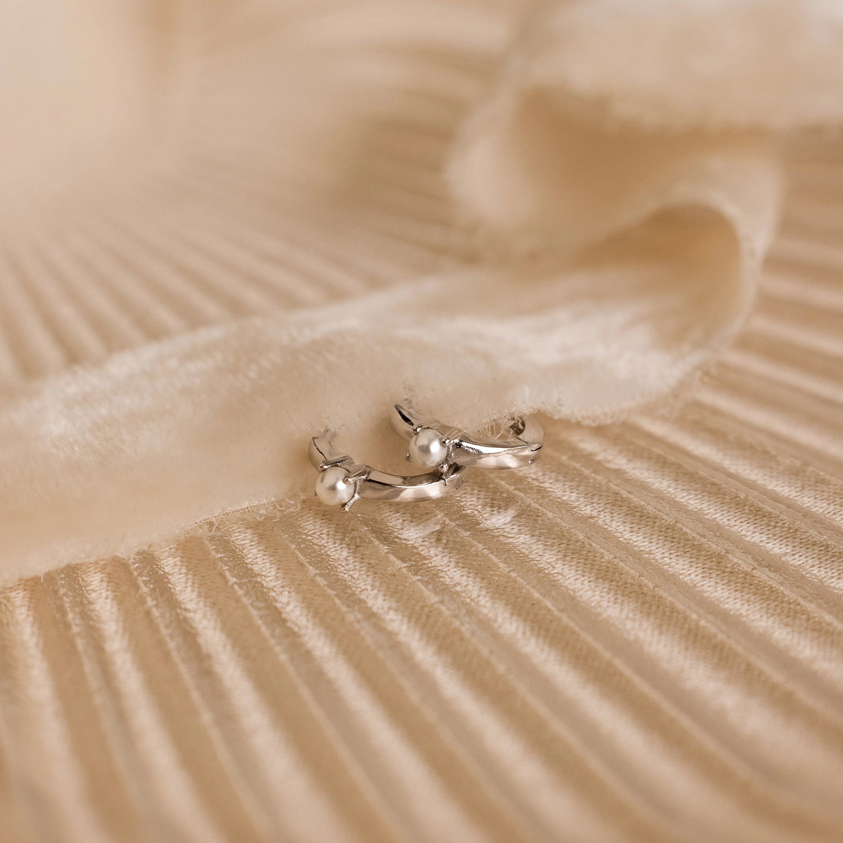 Tiny Pearl Huggies, featuring a delicate pearl accent, are displayed on a soft, cream-colored textured fabric background.