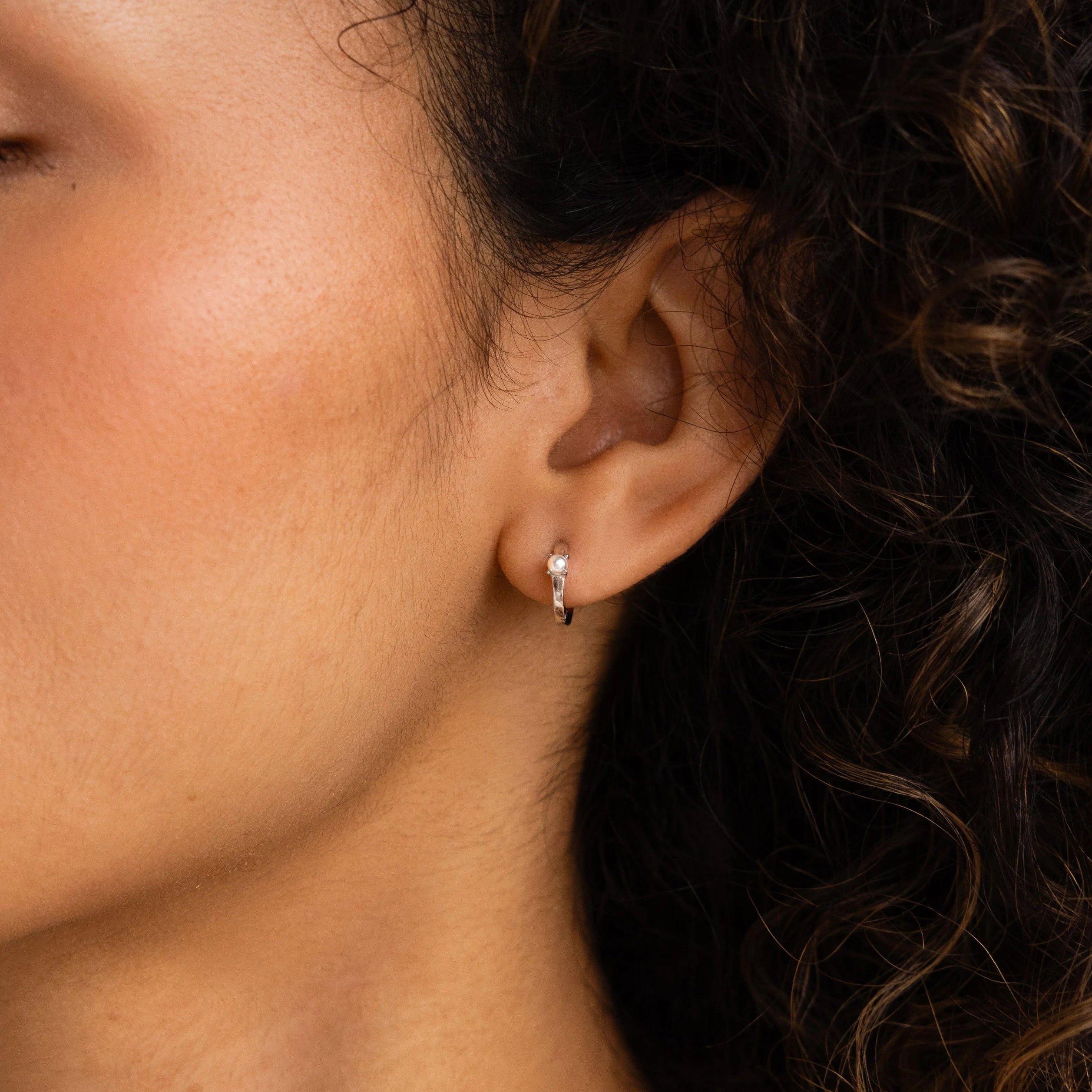 Close-up of a woman's ear wearing Tiny Pearl Huggies, chic hoops featuring a hint of pearl, with her curly hair partially visible in the frame.