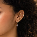 Close-up of a woman's ear adorned with multiple gold earrings, featuring Tiny Pearl Huggies alongside a pearl drop hoop and small studs.