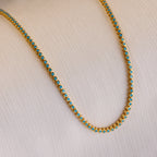 The Turquoise Bezel Tennis Necklace, featuring small turquoise stones set in gold, is displayed on a light textured fabric background.