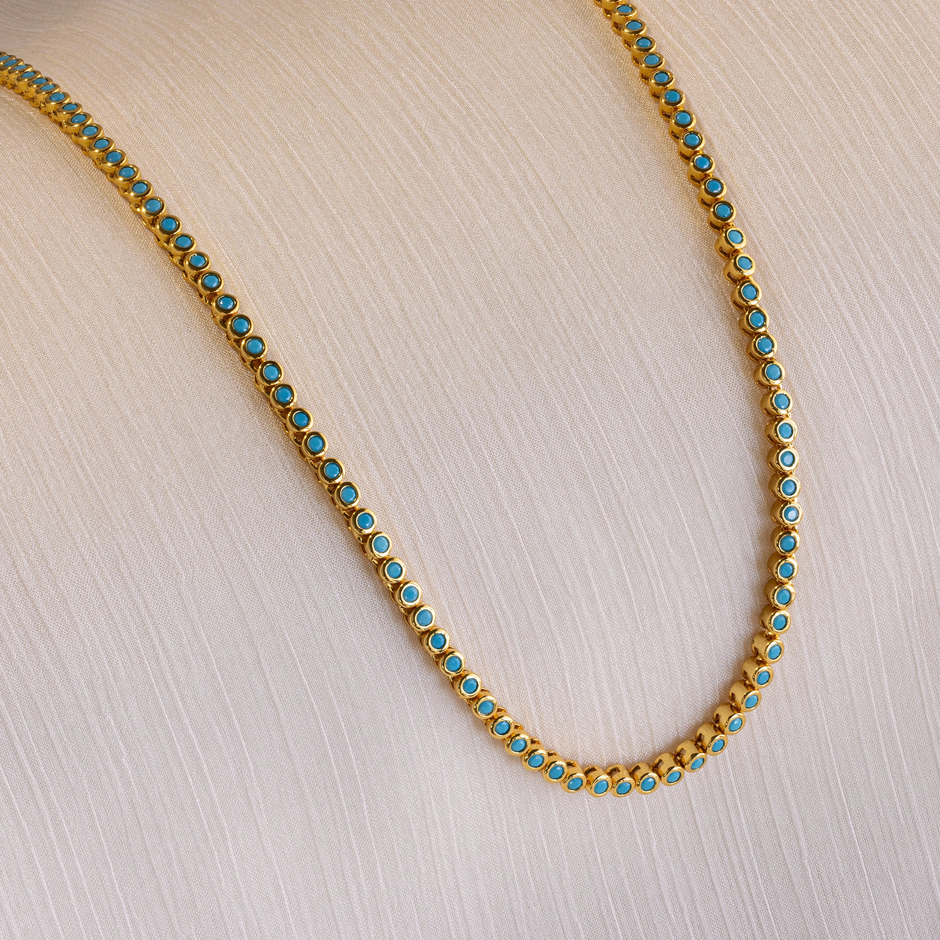 The Turquoise Bezel Tennis Necklace, featuring small turquoise stones set in gold, is displayed on a light textured fabric background.