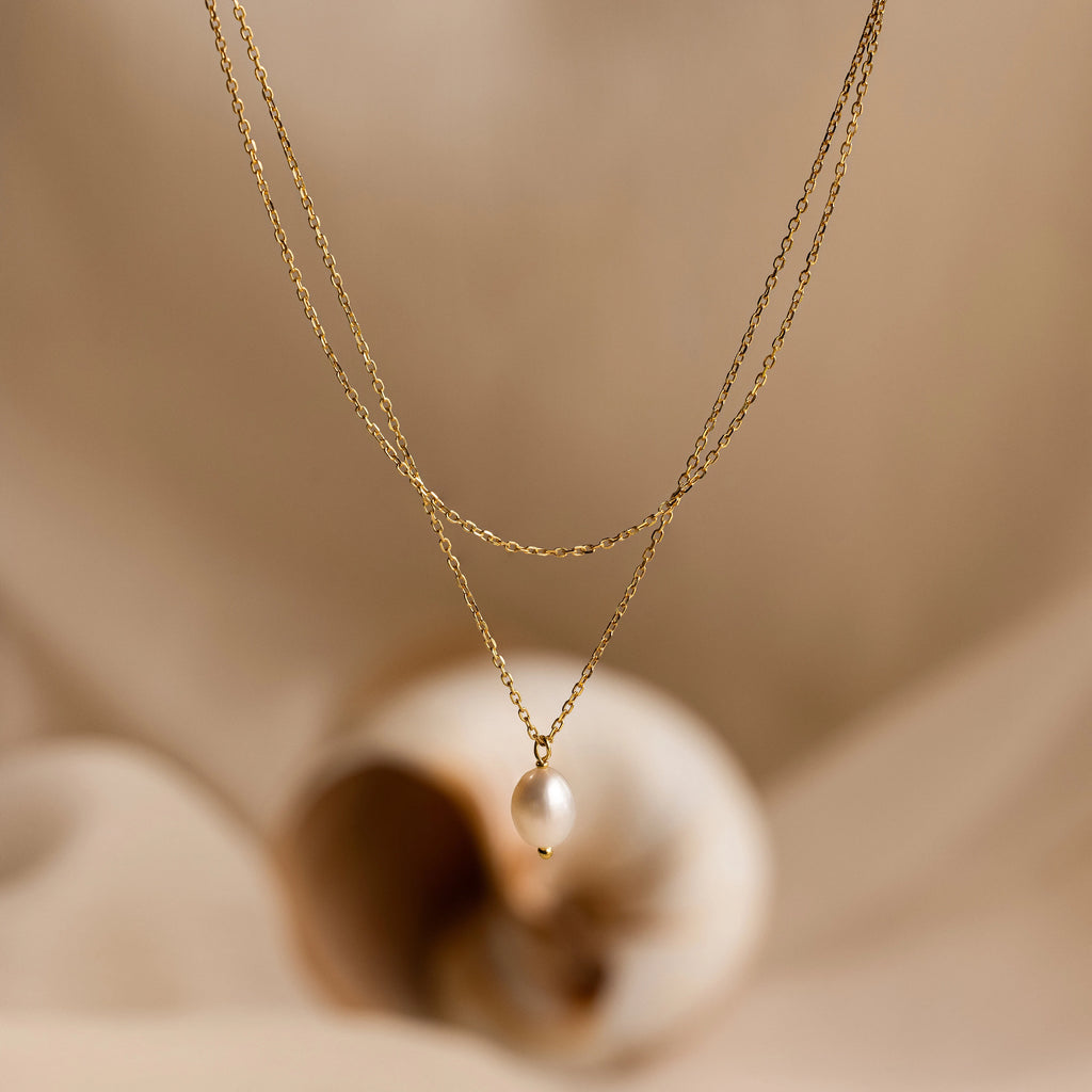 Duo Chain Pearl Necklace with a single pearl pendant, showcased on a soft beige background alongside a shell.