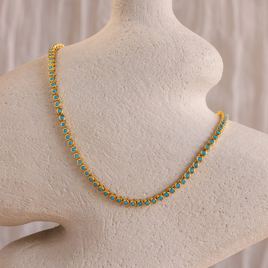 The Turquoise Bezel Tennis Necklace, featuring gold and turquoise stones, is displayed on a textured off-white mannequin bust.