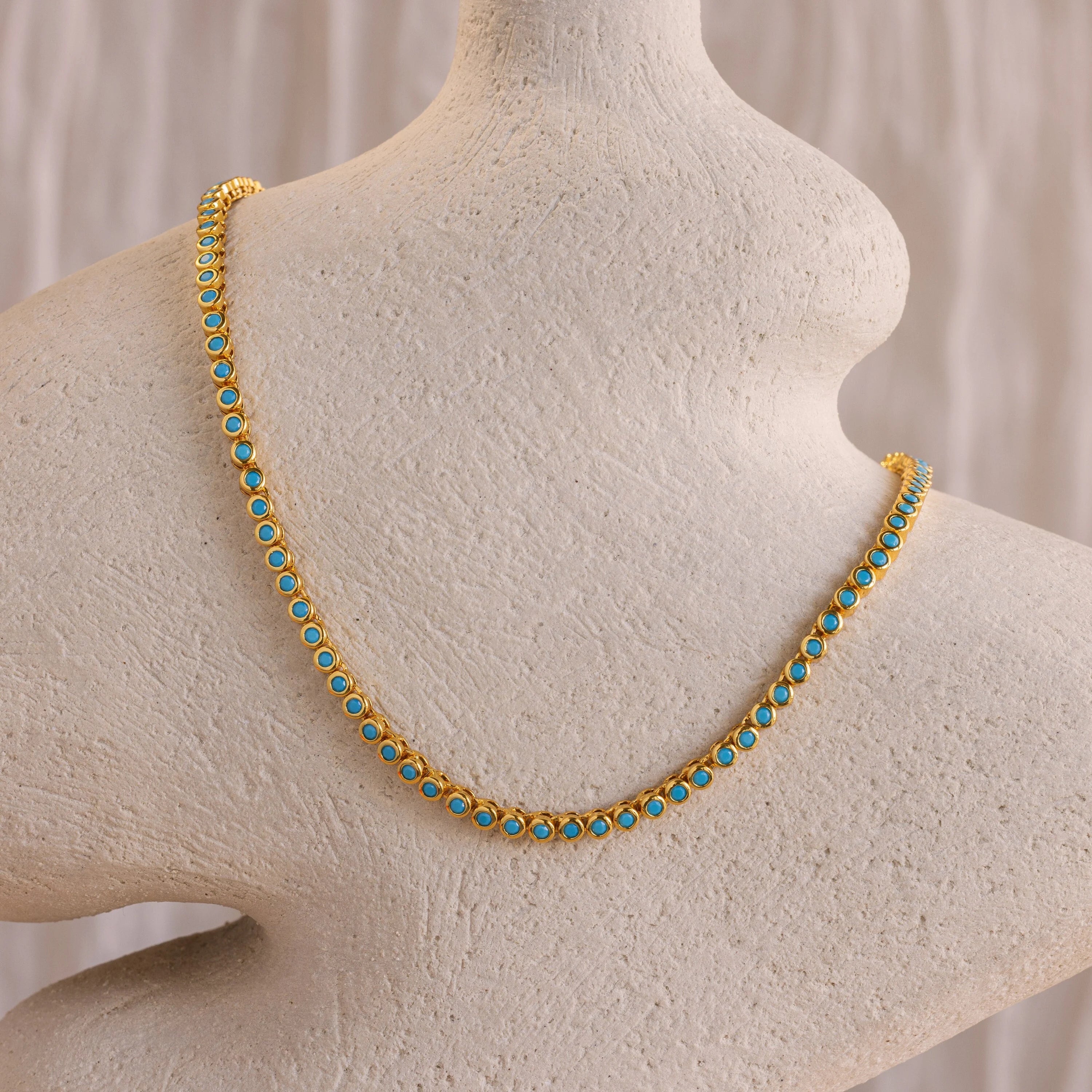 The Turquoise Bezel Tennis Necklace, featuring gold and turquoise stones, is displayed on a textured off-white mannequin bust.