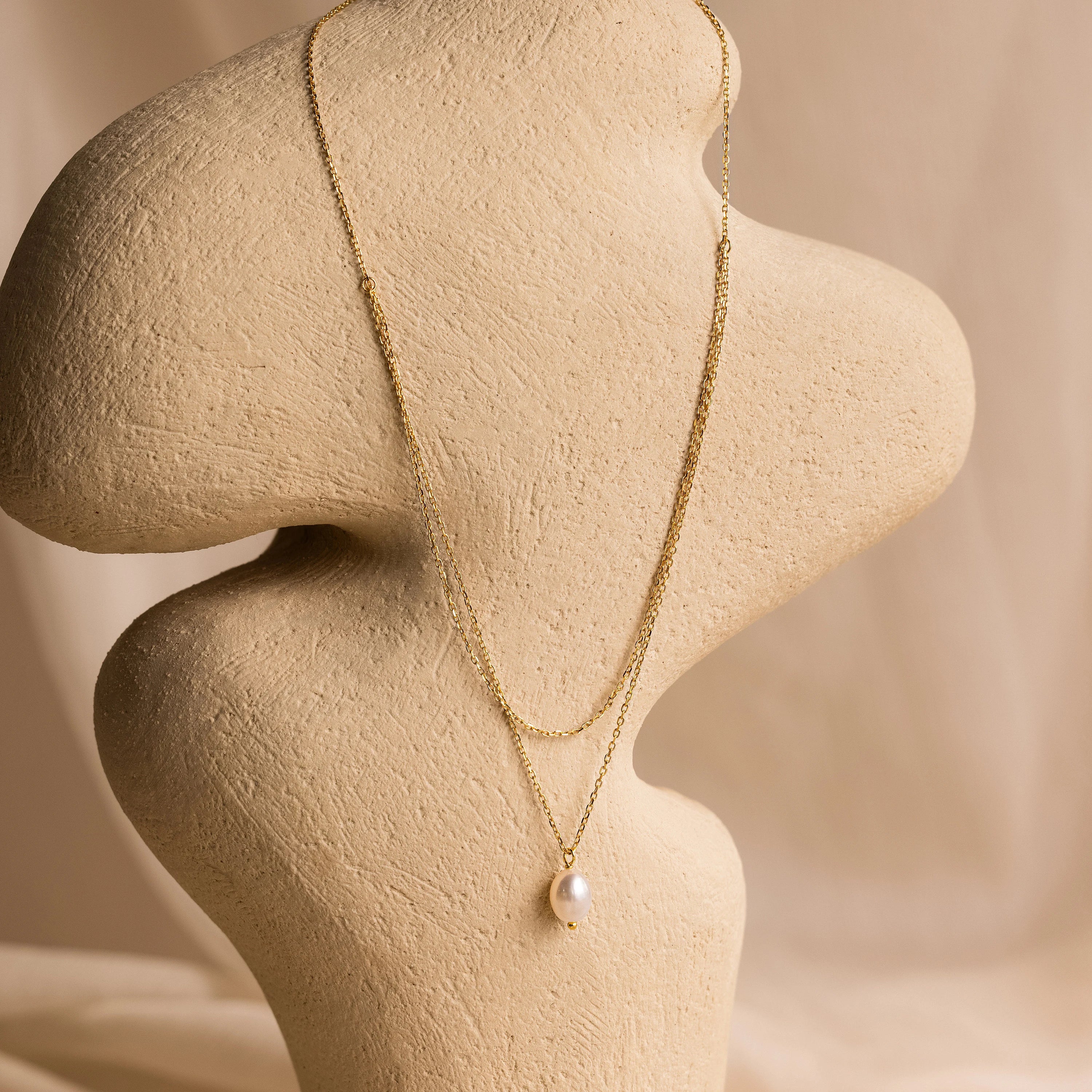 The Duo Chain Pearl Necklace features elegant pearls and a single pendant, showcased on a beige abstract bust with a soft neutral background. This timeless piece is perfect for wearing alone or layered with other necklaces.