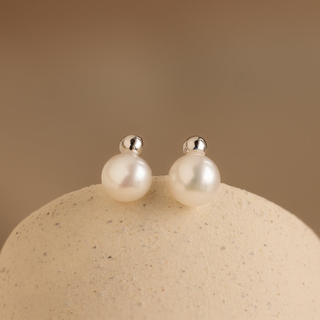 Classic Pearl Studs featuring silver posts are displayed on a smooth beige surface.