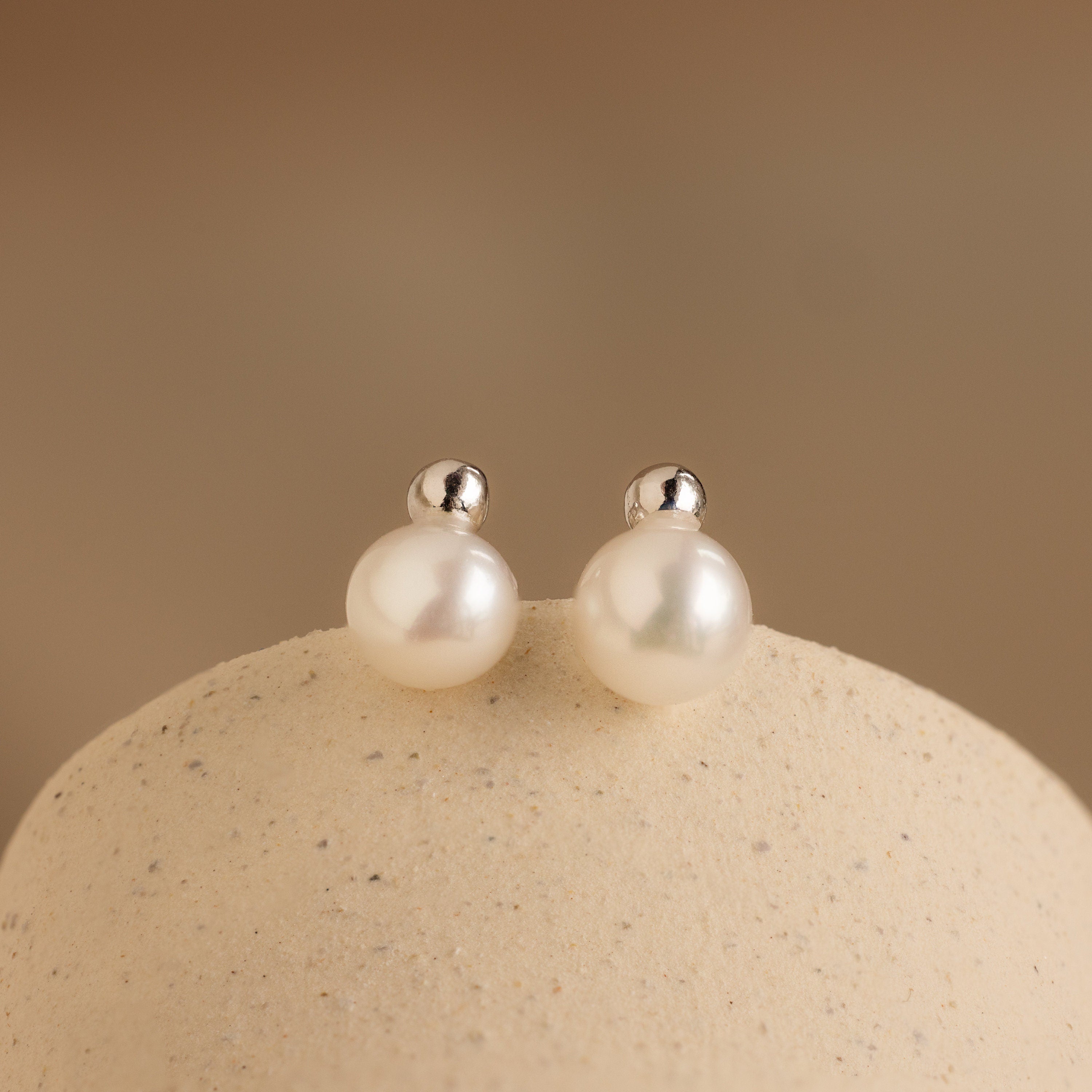Classic Pearl Studs featuring silver posts are displayed on a smooth beige surface.
