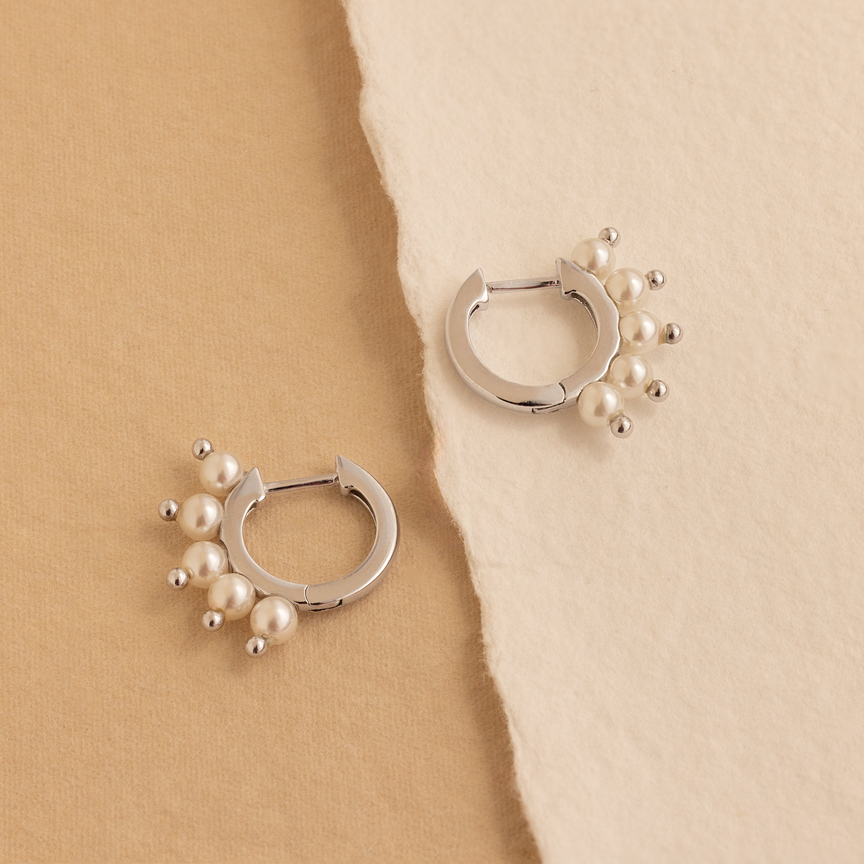 Two Beaded Pearl Huggies featuring clusters of small pearls are displayed on beige textured paper—a refined, modern take on pearl jewelry.