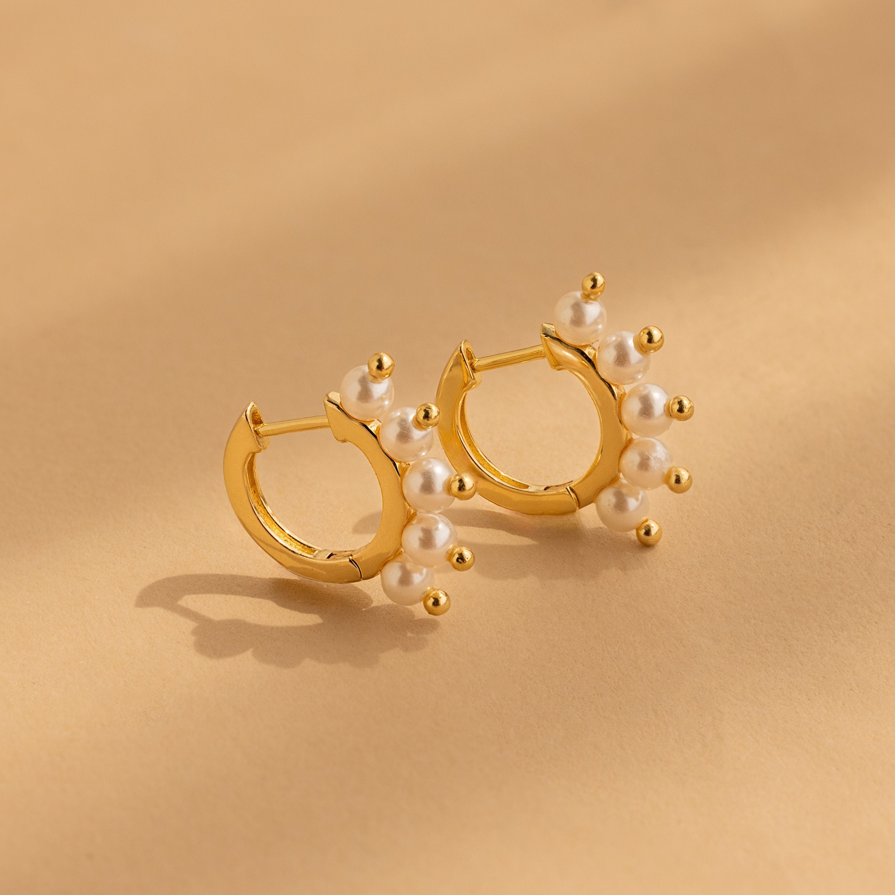 Beaded Pearl Huggies—gold hoop earrings with delicate white pearls—capture modern pearl jewelry style, shown here on a beige surface.