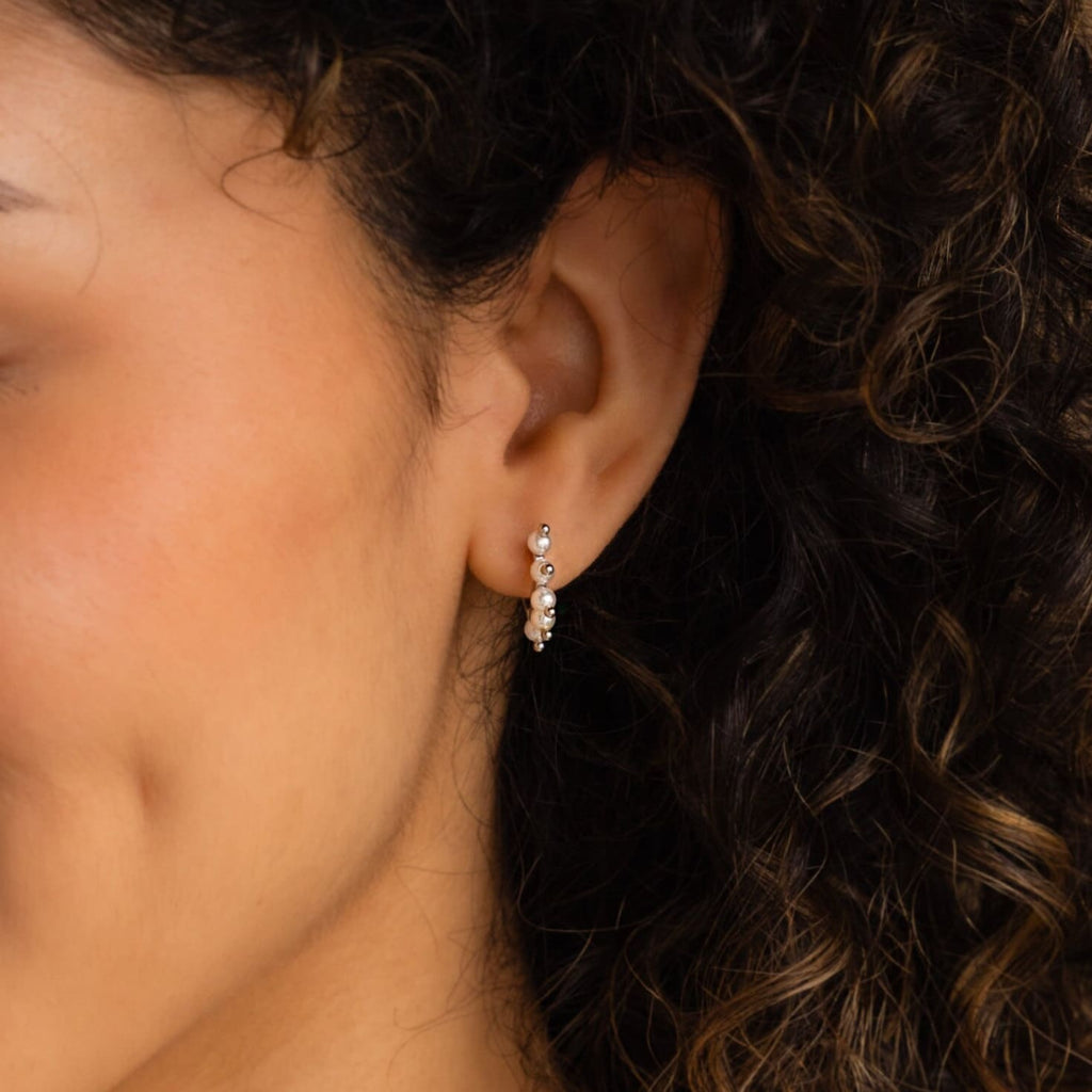 Close-up of a woman’s ear with curly hair, wearing Beaded Pearl Huggies—a modern pearl jewelry style featuring small silver hoops with round beaded details.