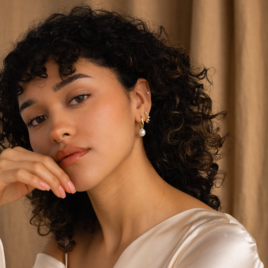 A woman with curly hair and a cream satin top poses gracefully, showcasing the Beaded Pearl Huggies modern pearl earrings with her hand near her face.