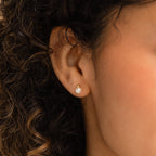 A woman’s ear is adorned with Classic Pearl Studs, her curly hair gently framing the timeless earring.