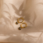 The Onyx Flower Huggies, gold hoop earrings adorned with small onyx flower charms, are displayed on soft beige fabric with gentle shadows.