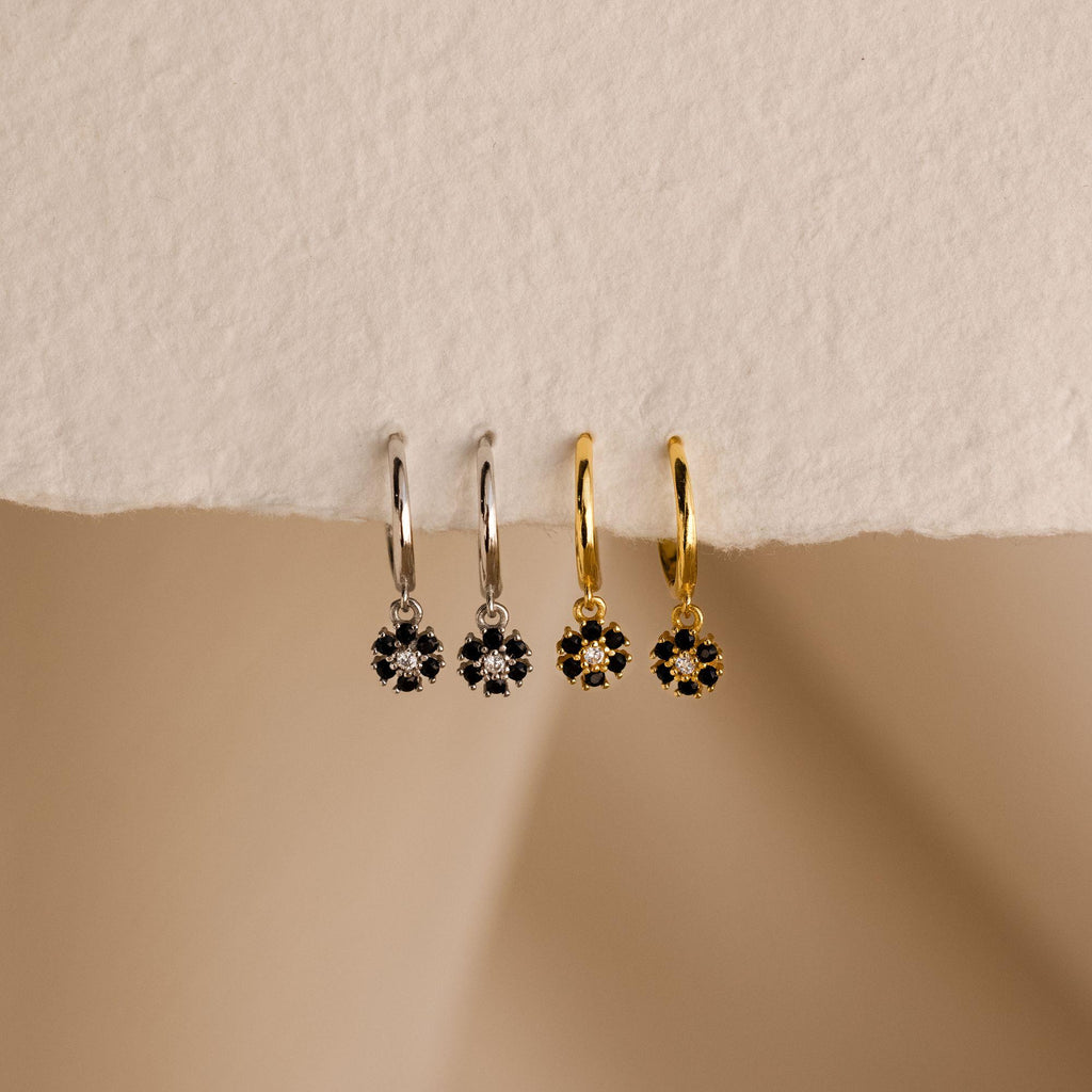 The Onyx Flower Huggies, in silver and gold with black onyx flower pendants, rest on a textured surface—perfect floral earrings for any occasion.