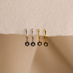 The Onyx Flower Huggies, in silver and gold with black onyx flower pendants, rest on a textured surface—perfect floral earrings for any occasion.