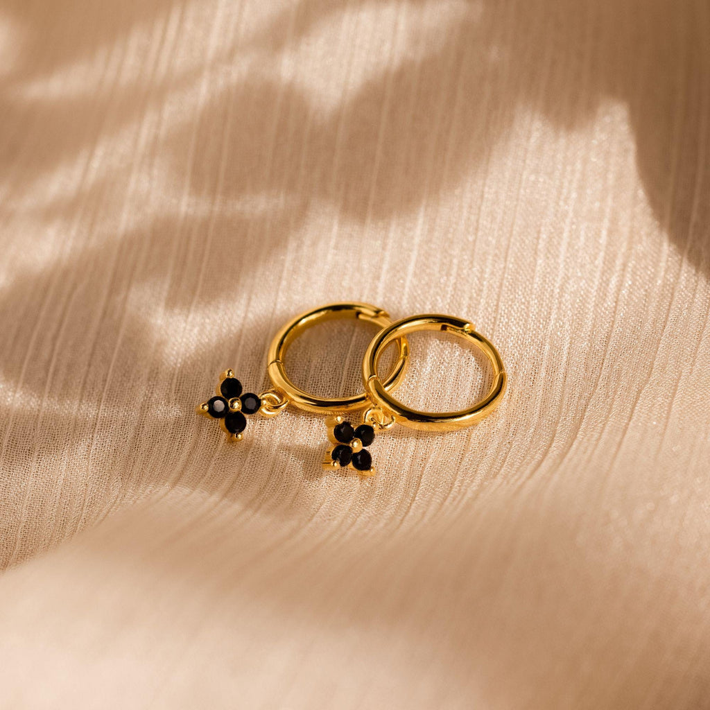 Black Ivy Flower Huggies—gold hoop earrings with black flower charms—rest on textured beige fabric, casting soft shadows.