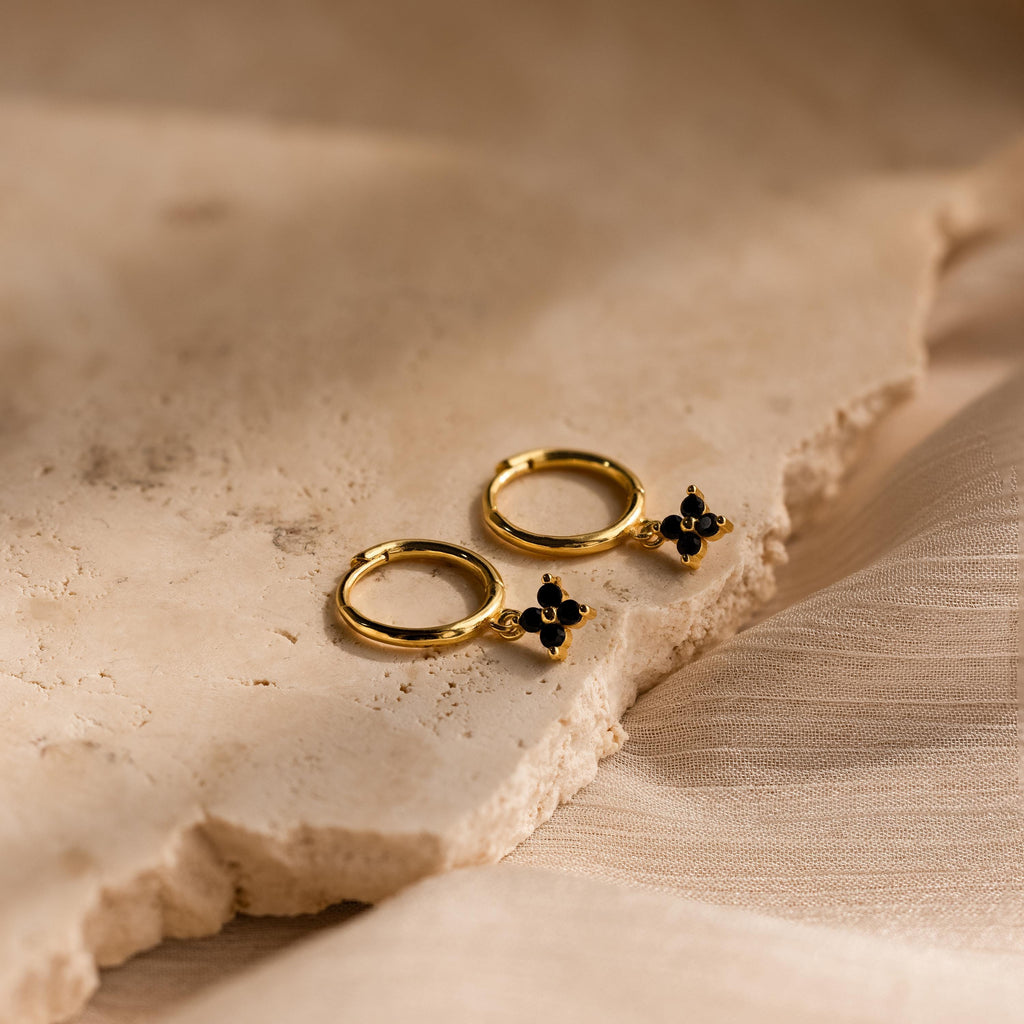 Black Ivy Flower Huggies: gold hoop earrings featuring black floral charms, shown on a textured beige stone surface.