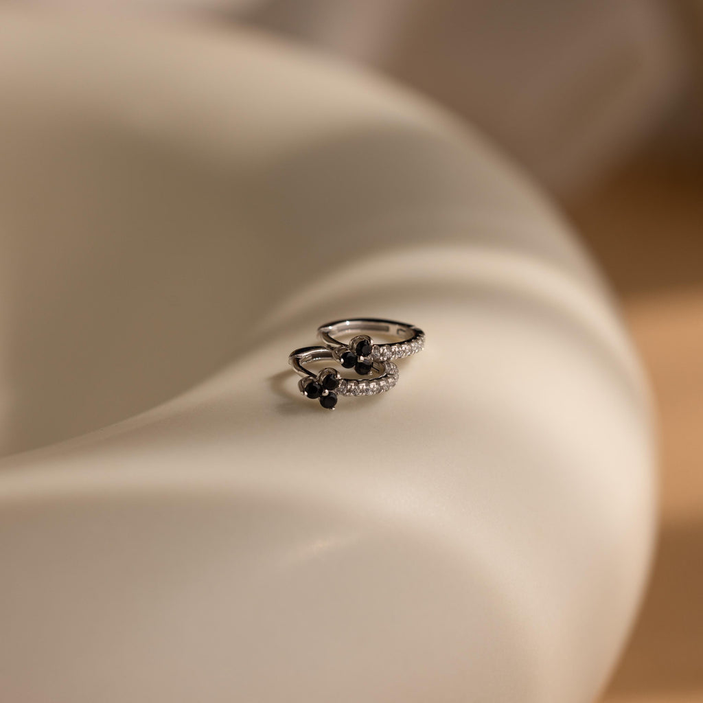 Two silver rings with black and clear stones rest on a smooth, curved, light surface, echoing the refined elegance of Onyx Lily Huggies.