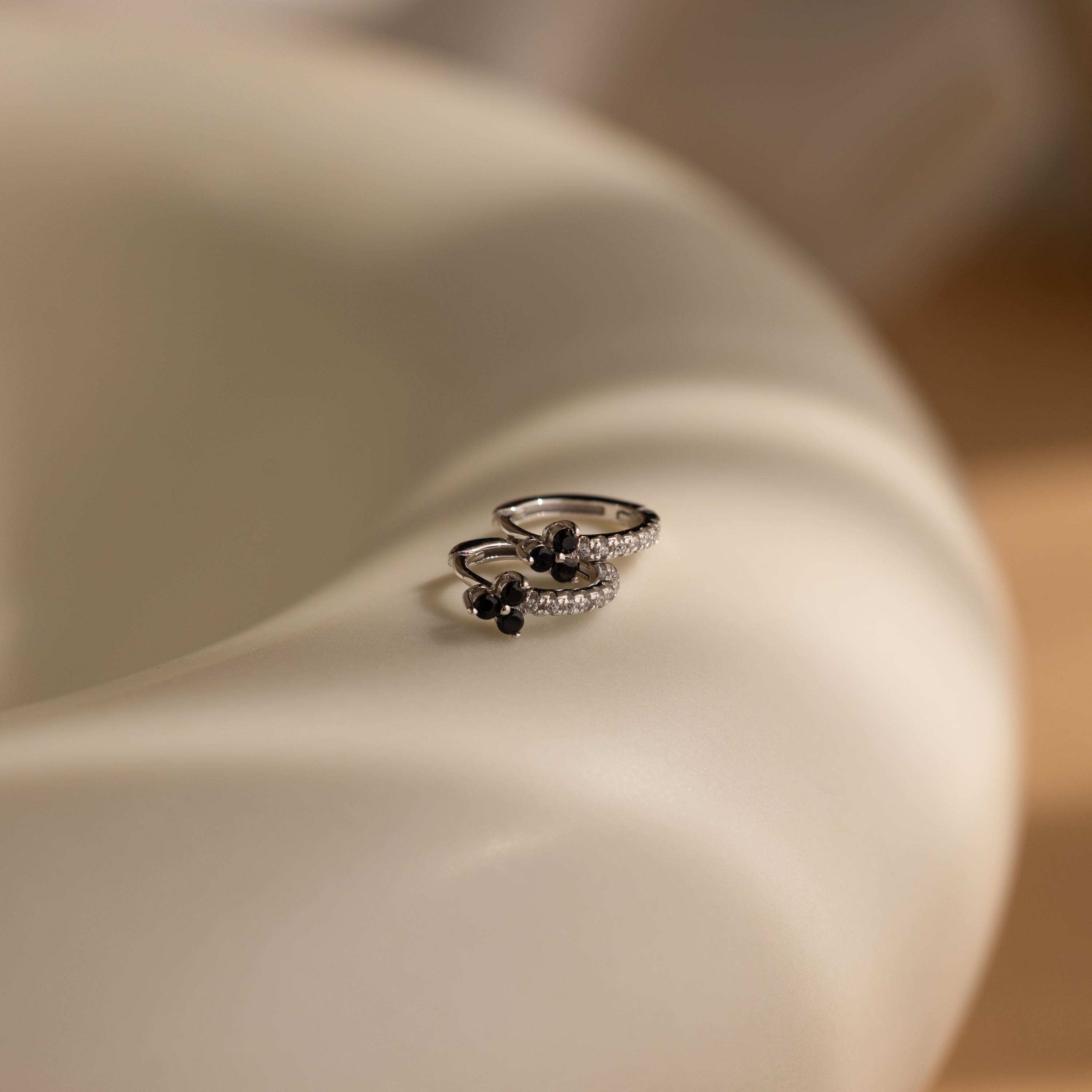 Two silver rings with black and clear stones rest on a smooth, curved, light surface, echoing the refined elegance of Onyx Lily Huggies.