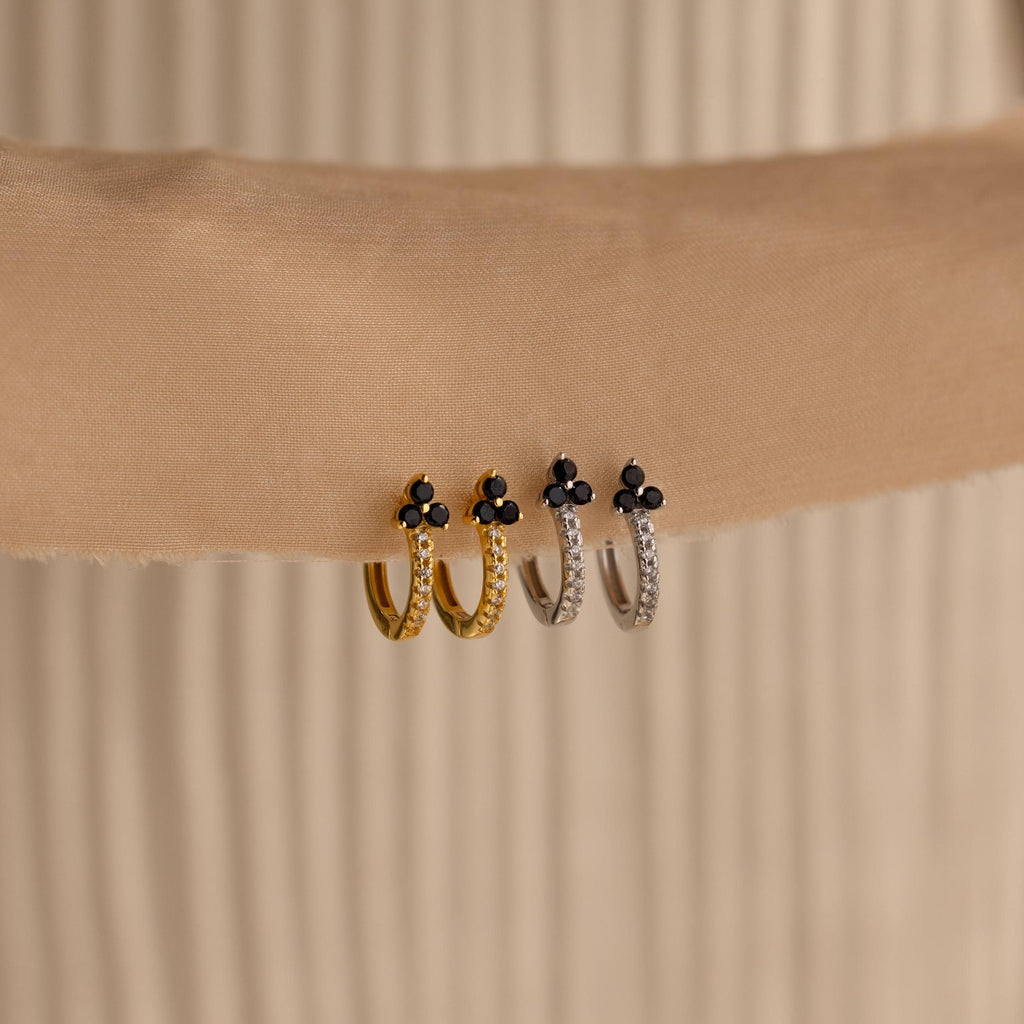 Two gold and two silver Onyx Lily Huggies, featuring small black stones, are displayed on a beige fabric background.