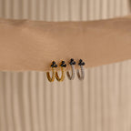 Two gold and two silver Onyx Lily Huggies, featuring small black stones, are displayed on a beige fabric background.