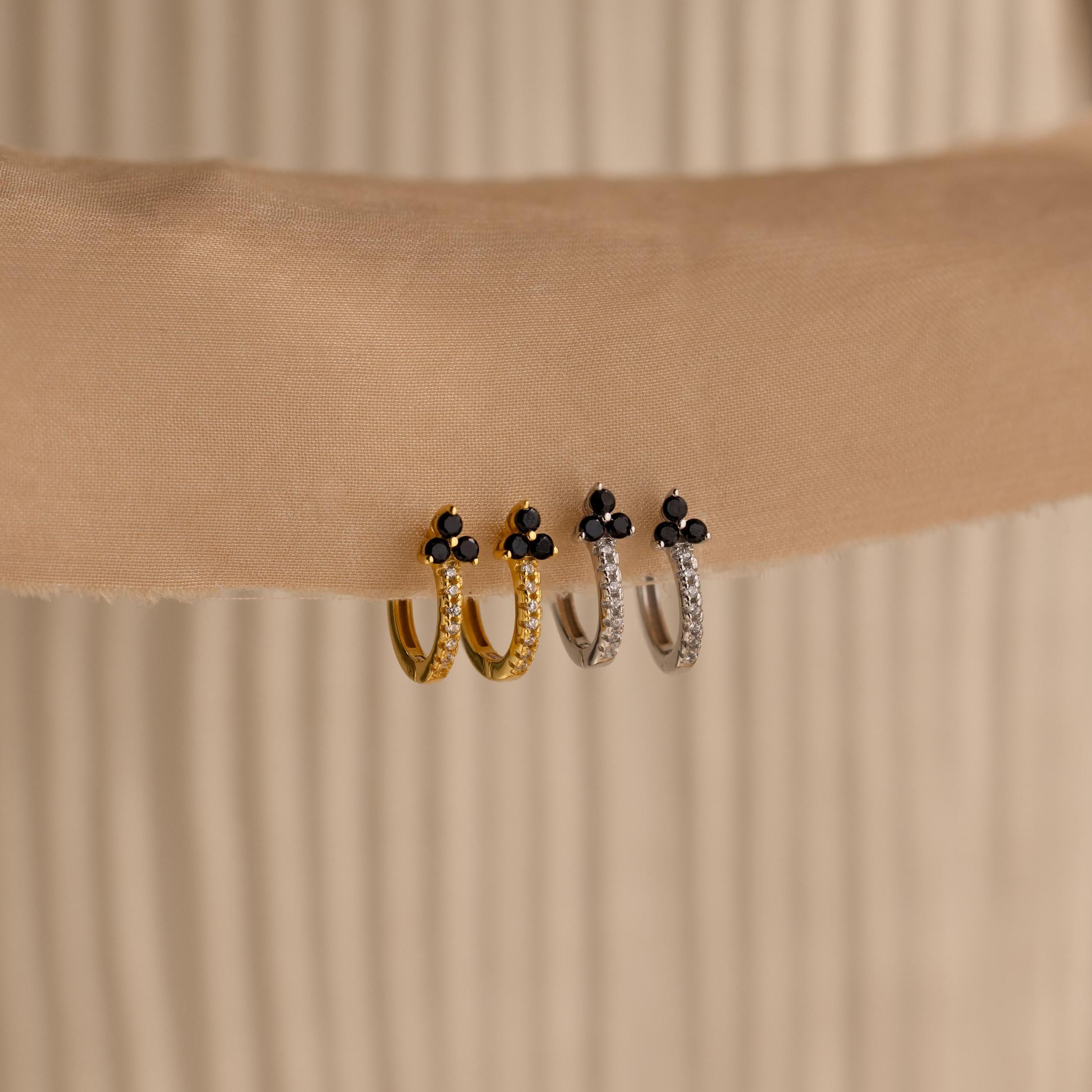 Two gold and two silver Onyx Lily Huggies, featuring small black stones, are displayed on a beige fabric background.