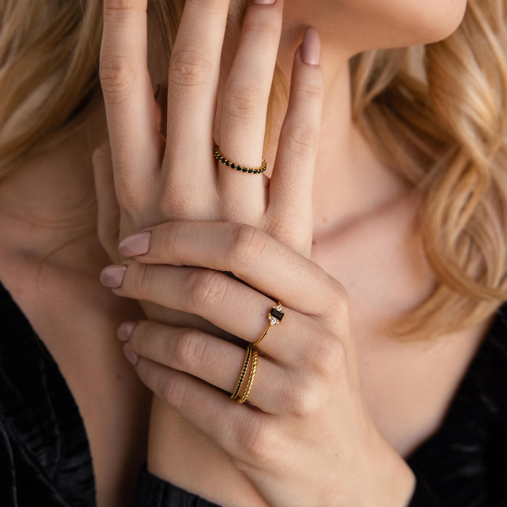A woman with manicured nails displays several gold rings, including the bold Black Baguette Diamond Ring in 18K Gold, worn as a statement piece on her fingers in this close-up shot.