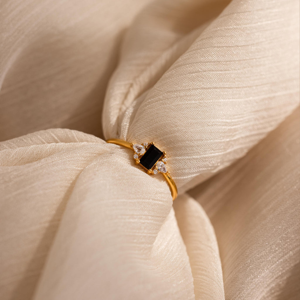 The Black Baguette Diamond Ring in 18K Gold features a rectangular black stone with clear gem accents, showcased on textured cream fabric—a bold yet subtly vintage statement piece.