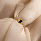 The Black Baguette Diamond Ring in 18K Gold features a rectangular black stone with clear gem accents, showcased on textured cream fabric—a bold yet subtly vintage statement piece.