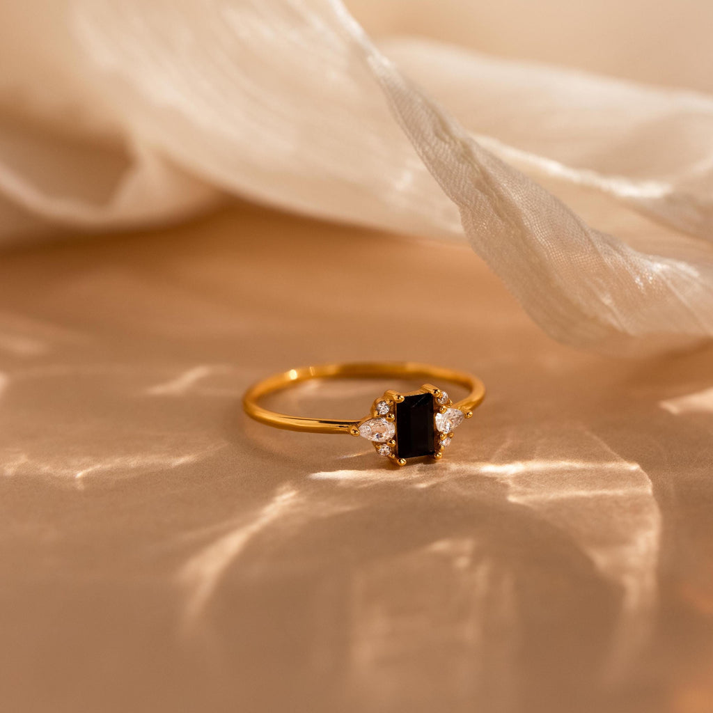 The Black Baguette Diamond Ring features a vintage-inspired gold band with a rectangular black stone and small clear gems, displayed on soft beige fabric.