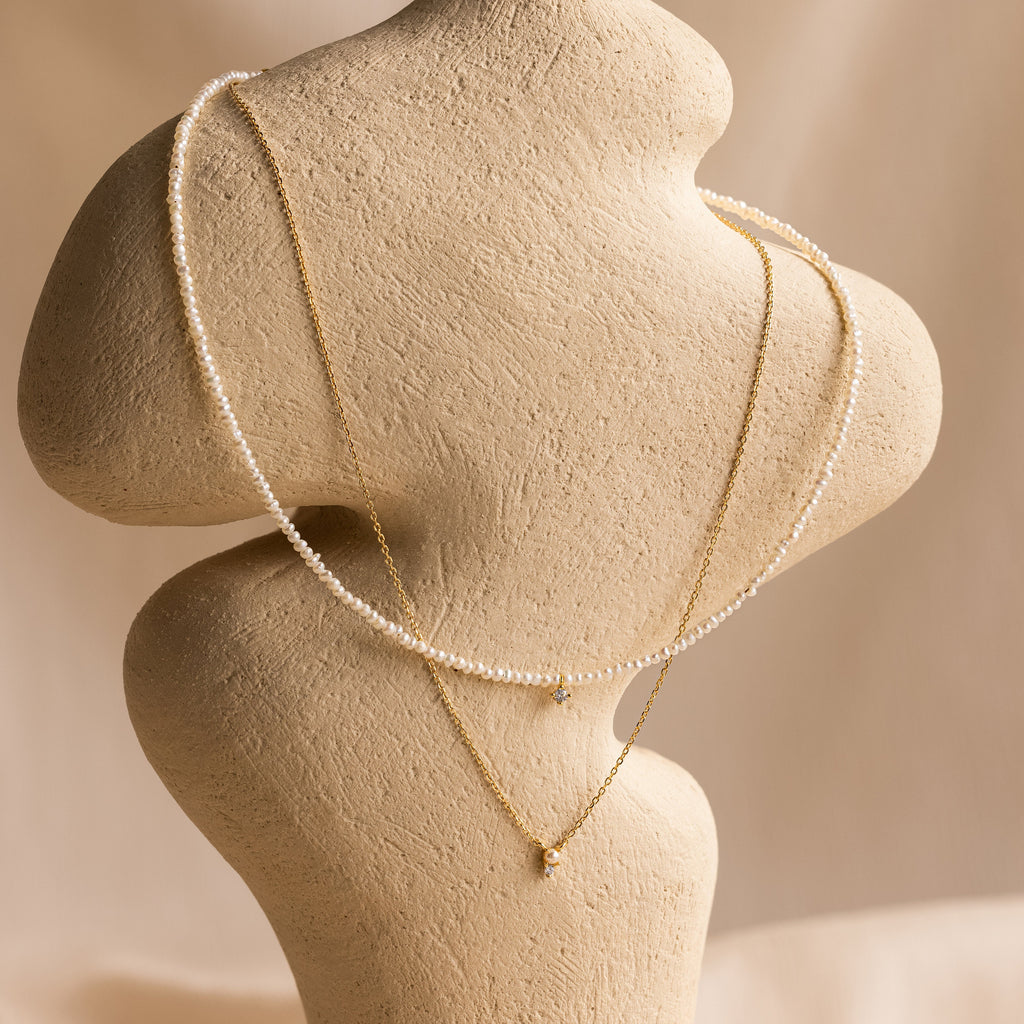 The Pearl & Diamond Duo Necklace is elegantly displayed on a textured beige mannequin against a soft, neutral background.
