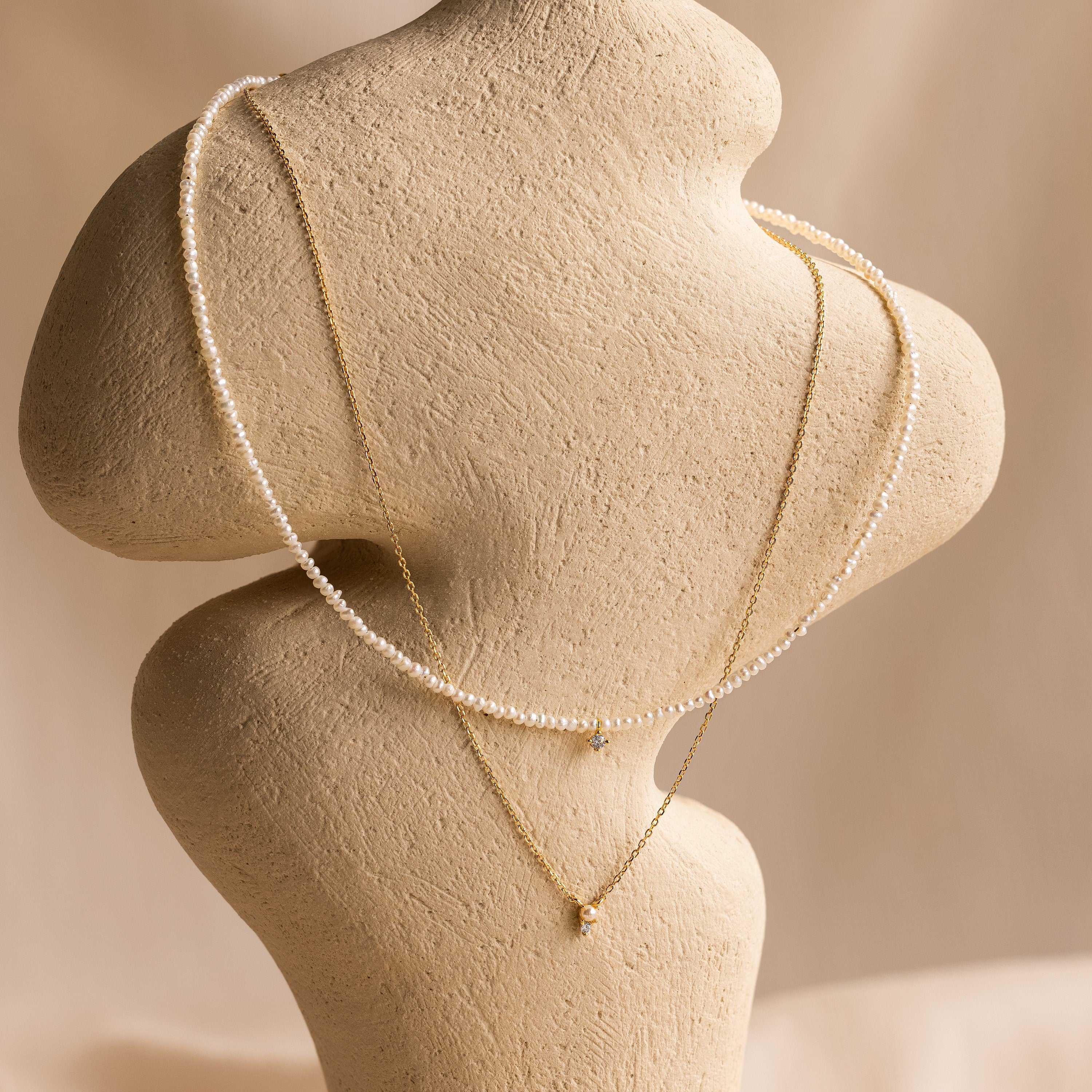 The Pearl & Diamond Duo Necklace is elegantly displayed on a textured beige mannequin against a soft, neutral background.