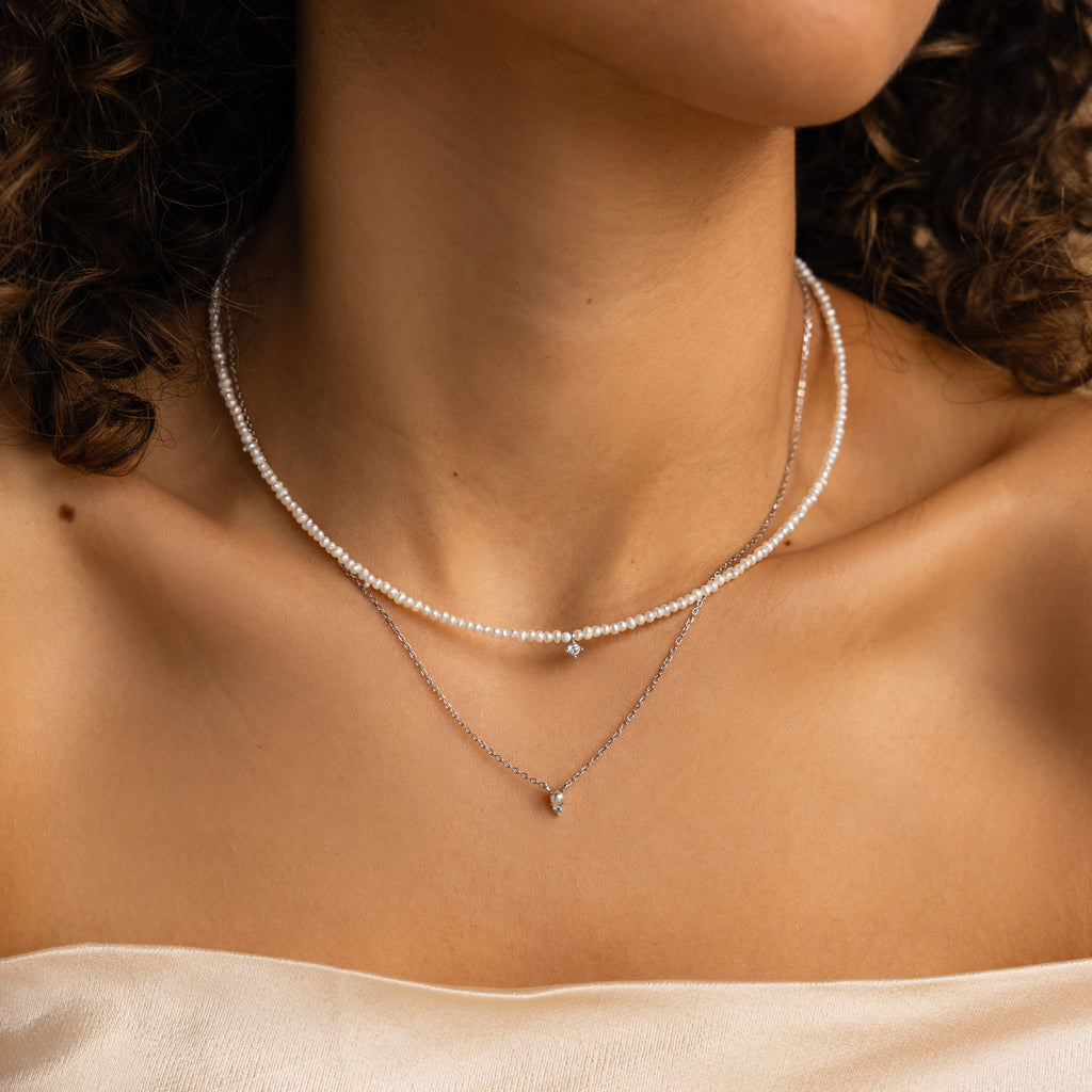 A woman wears the Pearl & Diamond Duo Necklace—two delicate layers featuring a pearl choker and a diamond pendant—gracefully draped against her bare shoulders.