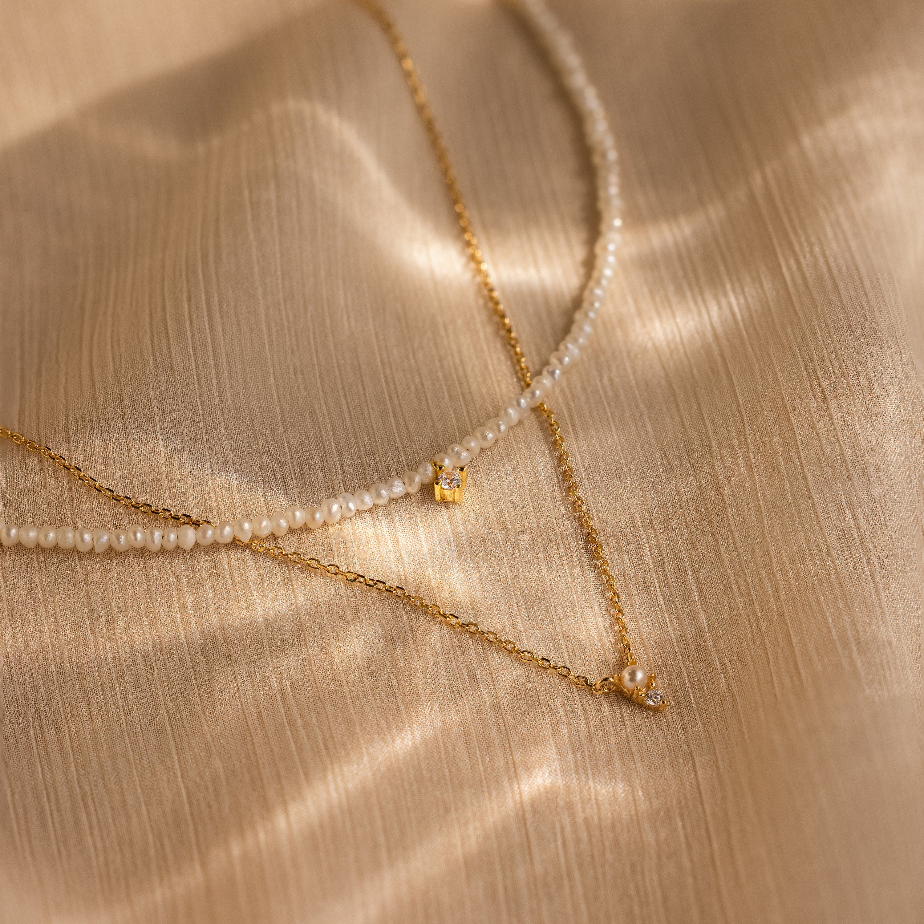 The Pearl & Diamond Duo Necklace, a delicate gold piece, is displayed on a textured beige fabric background.