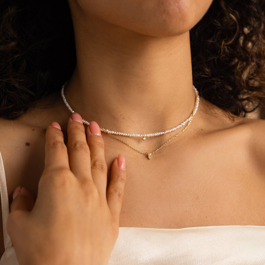 A woman in a white top touches her collarbone while wearing layered gold and pearl necklaces, featuring the Pearl & Diamond Duo Necklace.