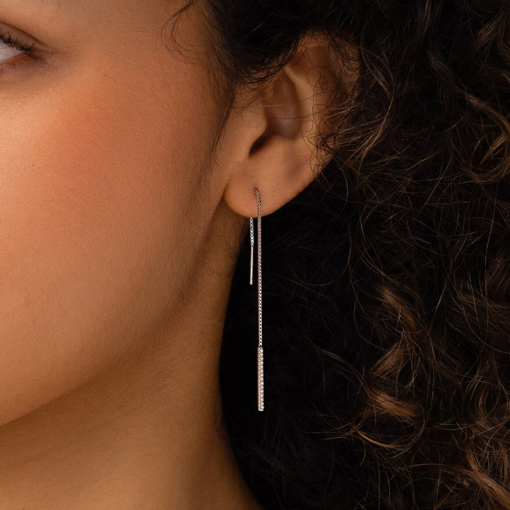 A close-up shows a woman's ear wearing Pearl Bar Threader Earrings, featuring delicate silver strands and dainty pearls that dangle elegantly against her curly hair.
