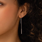 A close-up shows a woman's ear wearing Pearl Bar Threader Earrings, featuring delicate silver strands and dainty pearls that dangle elegantly against her curly hair.
