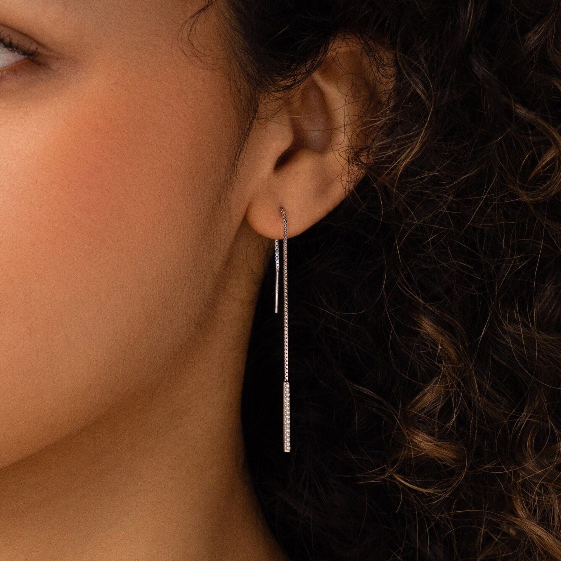 A close-up shows a woman's ear wearing Pearl Bar Threader Earrings, featuring delicate silver strands and dainty pearls that dangle elegantly against her curly hair.