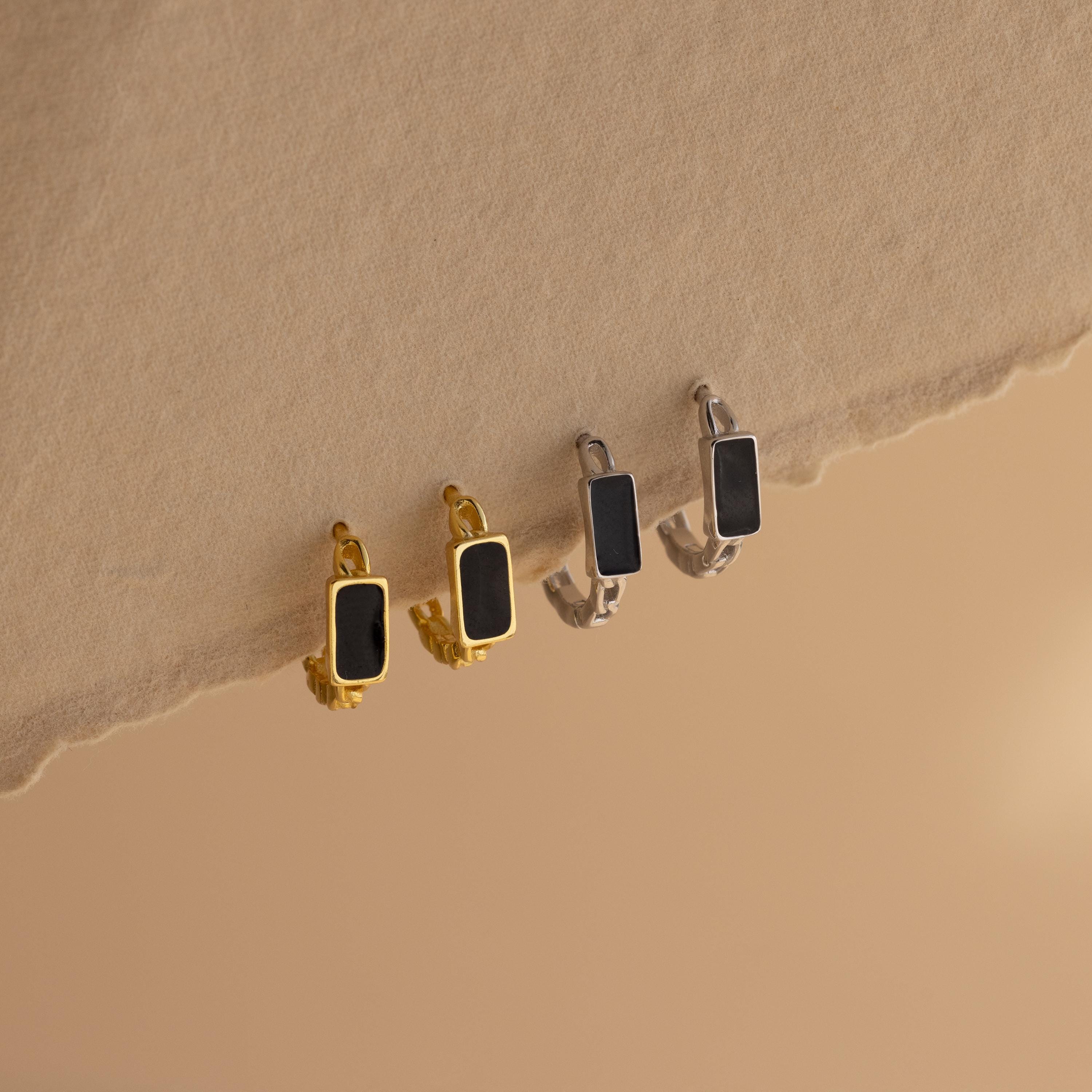 Two pairs of Onyx Link Huggies—one gold, one silver—featuring rectangular black onyx gemstones, are displayed on a textured beige surface. These bold earrings add modern elegance to any collection.