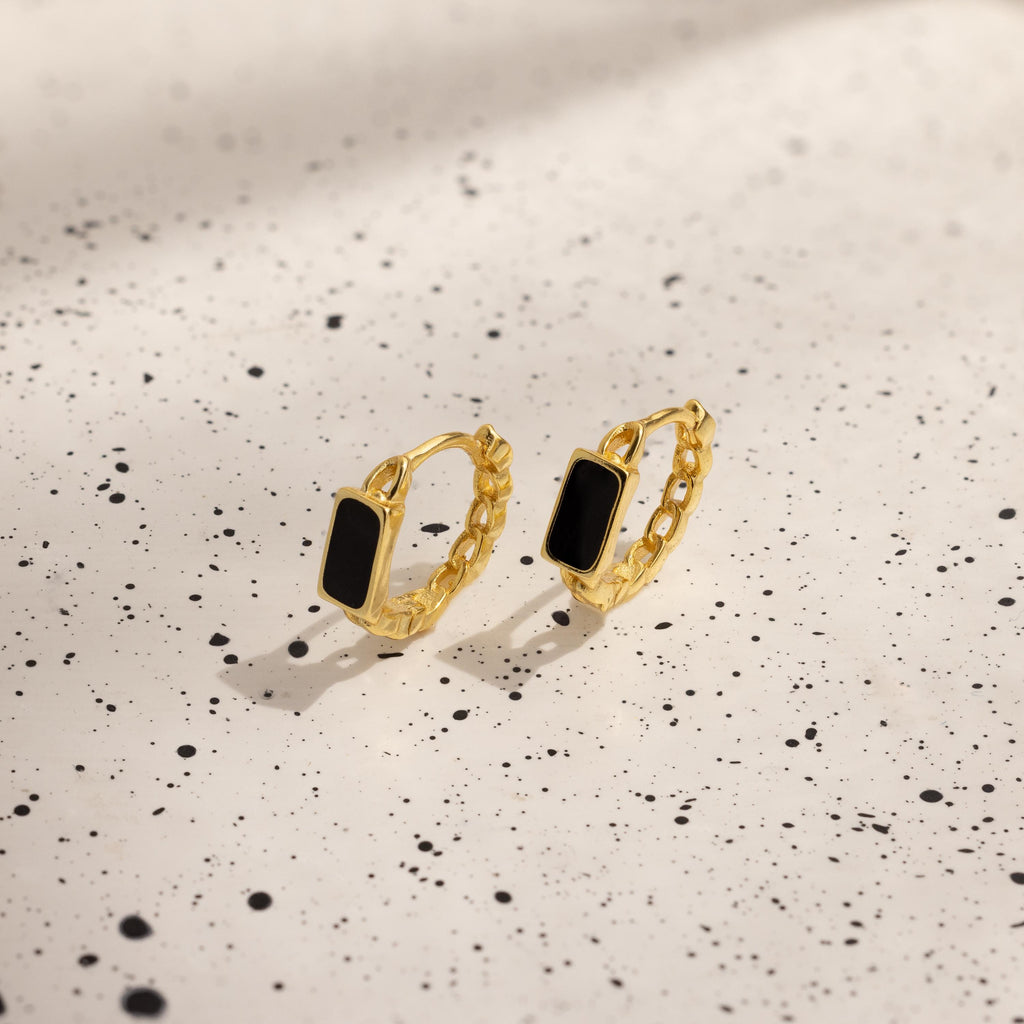 The Onyx Link Huggies, featuring gold chain hoops with black rectangular onyx gemstones, rest on a white speckled surface.