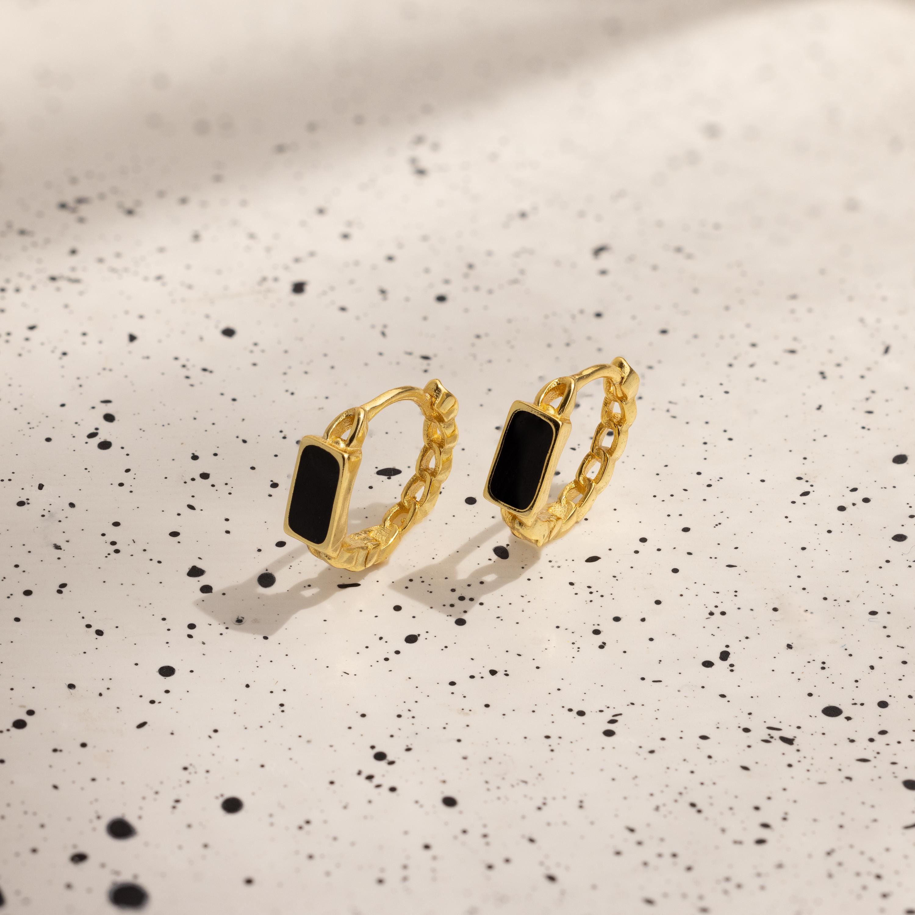 The Onyx Link Huggies, featuring gold chain hoops with black rectangular onyx gemstones, rest on a white speckled surface.