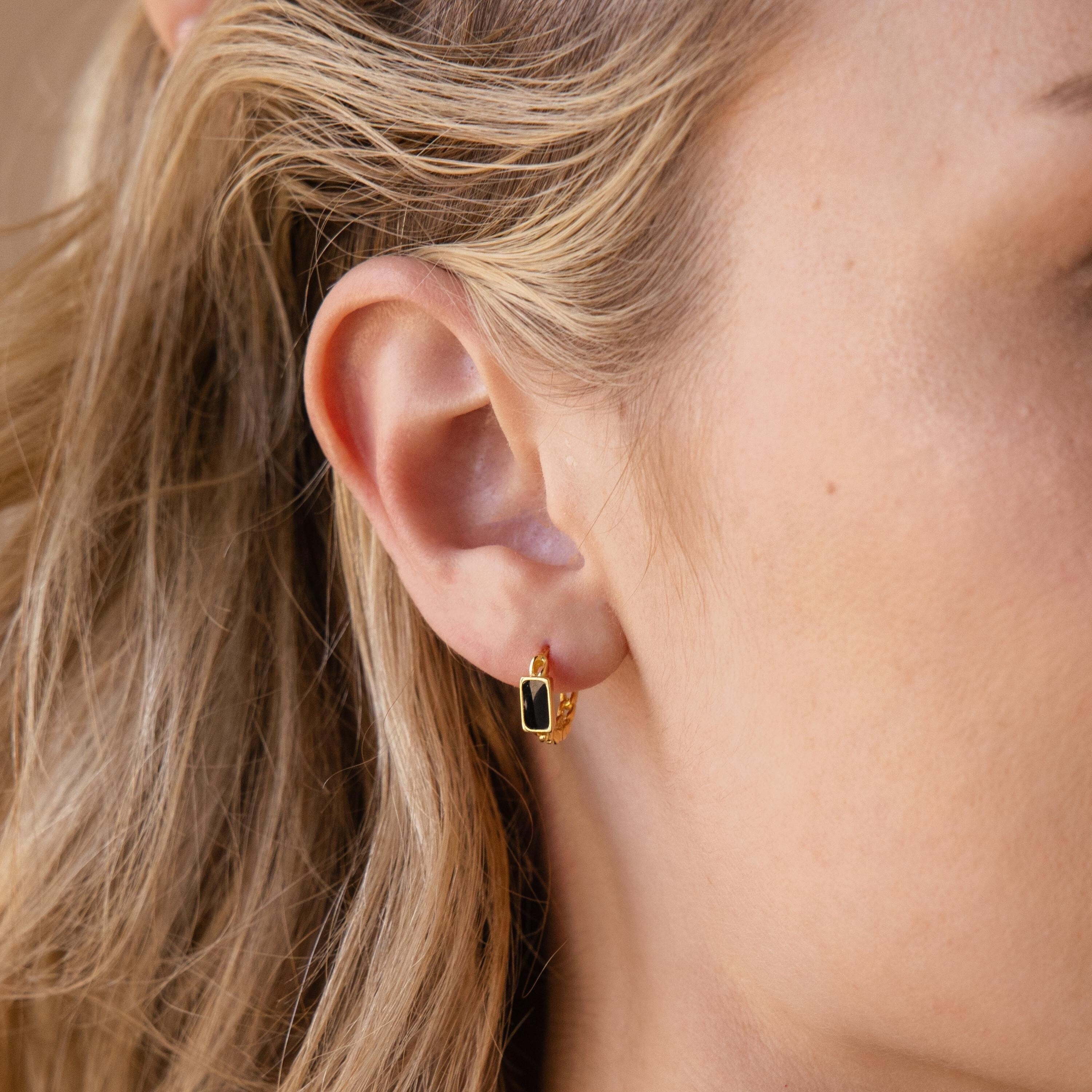 Close-up of a woman's ear wearing Onyx Link Huggies—chain hoop earrings adorned with a sleek rectangular black charm inspired by the elegance of onyx gemstones.
