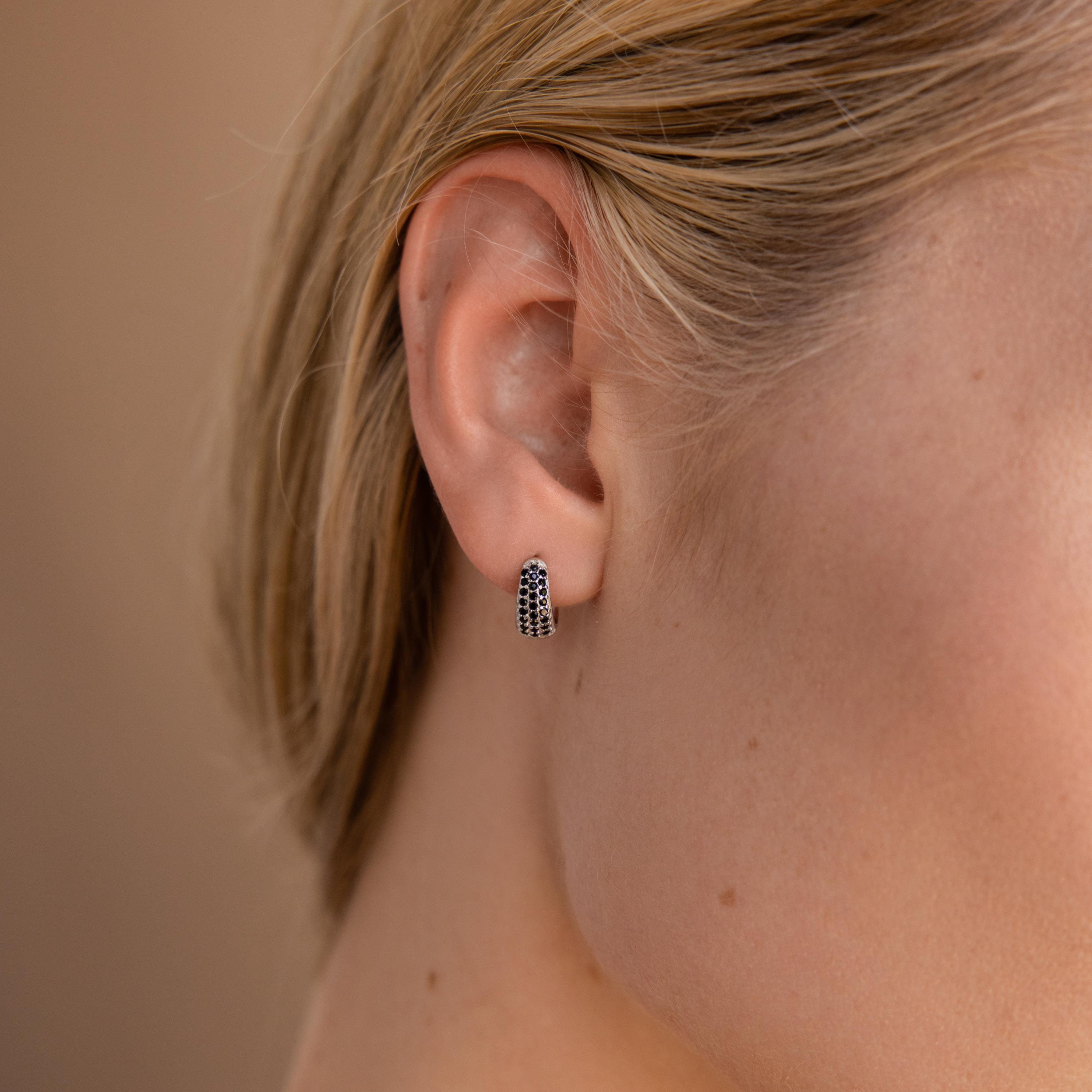 Close-up of a woman's ear wearing Chunky Onyx Pave Huggies—small silver hoops with black stones—against a light skin tone background.