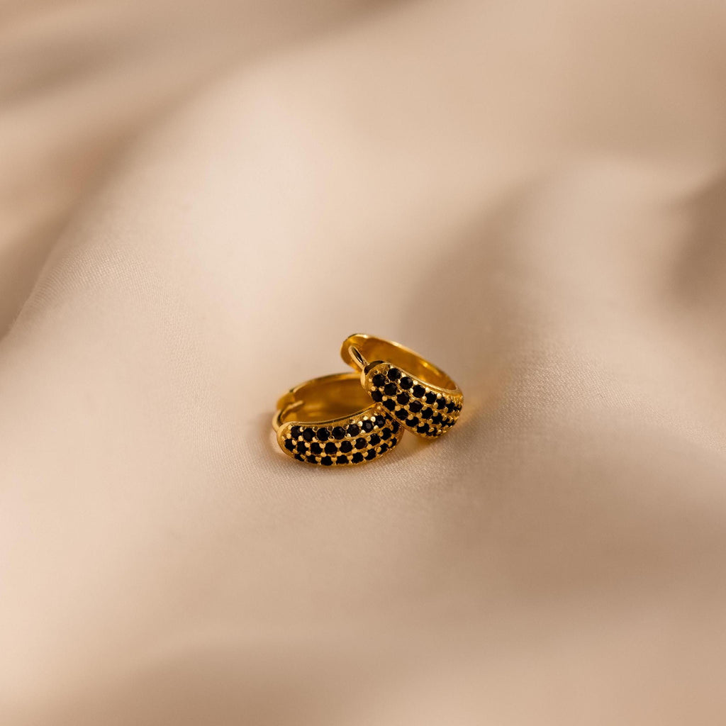 The Chunky Onyx Pave Huggies, a pair of gold modern hoops with black stones, rest on soft beige fabric—adding a chic touch to any look.