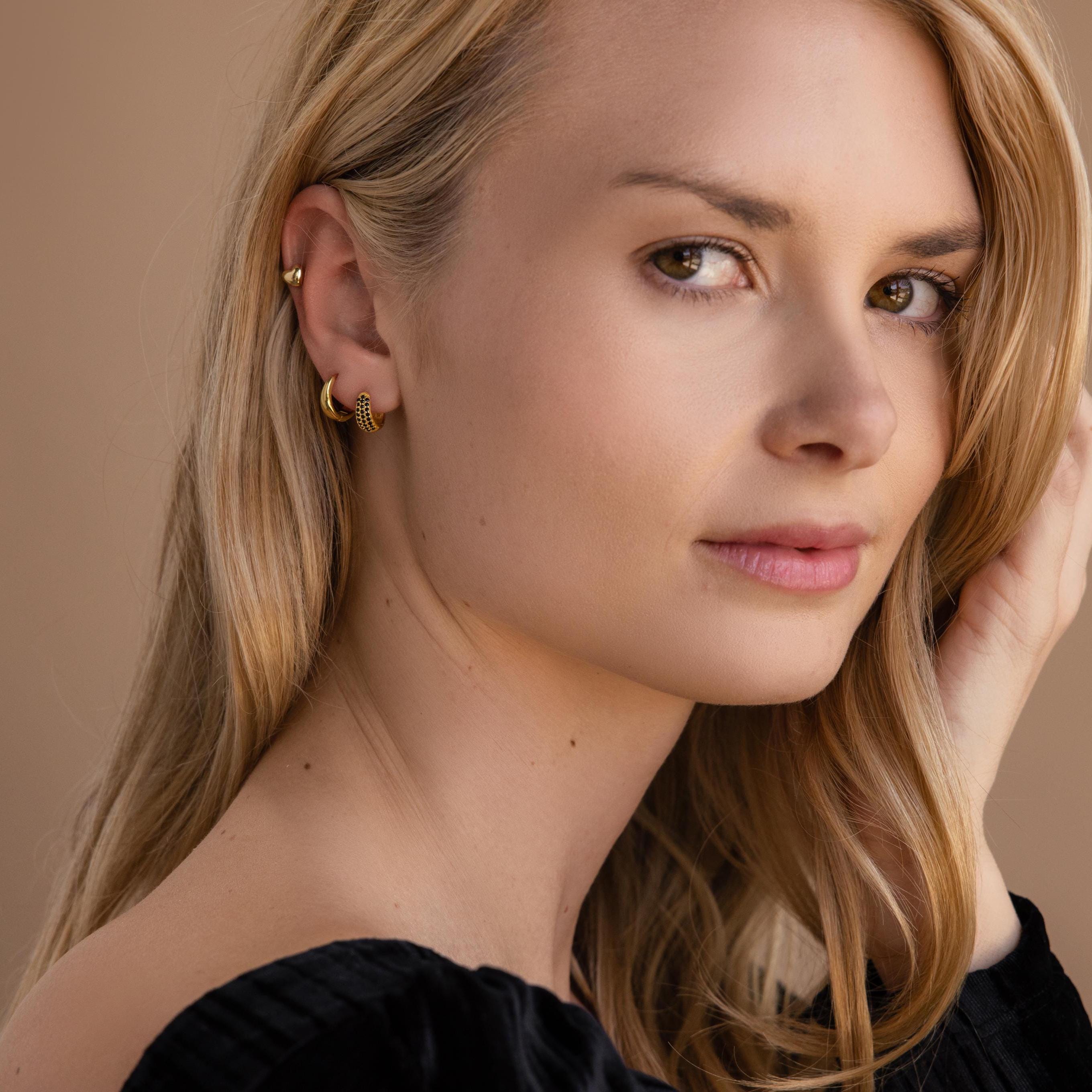 A blonde woman in a black top gazes at the camera, wearing Chunky Onyx Pave Huggies—modern hoops featuring small gold accents.