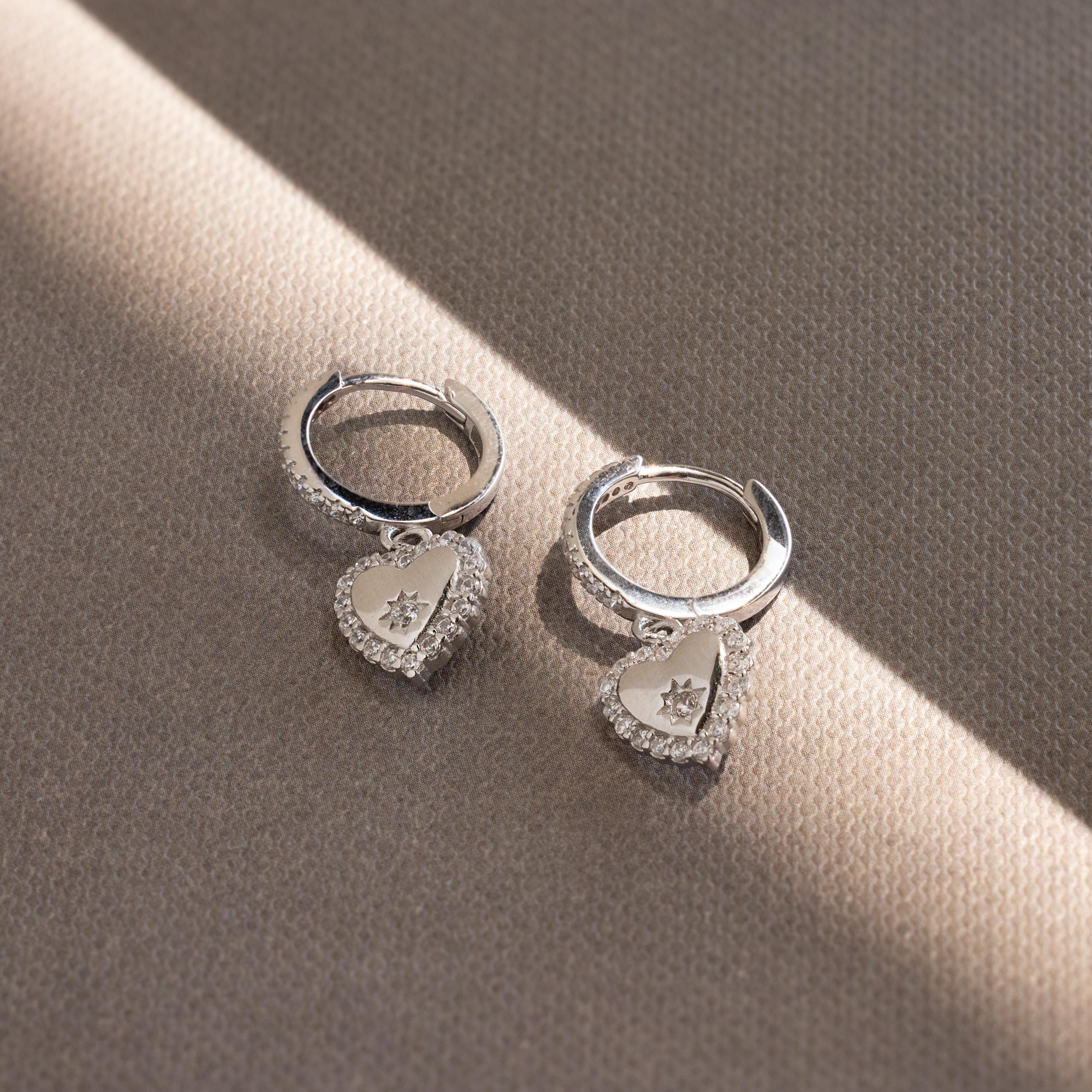 Pave Heart Drop Huggies featuring heart-shaped pendants and sparkling central stones rest on a textured beige surface—perfect as a romantic gift.